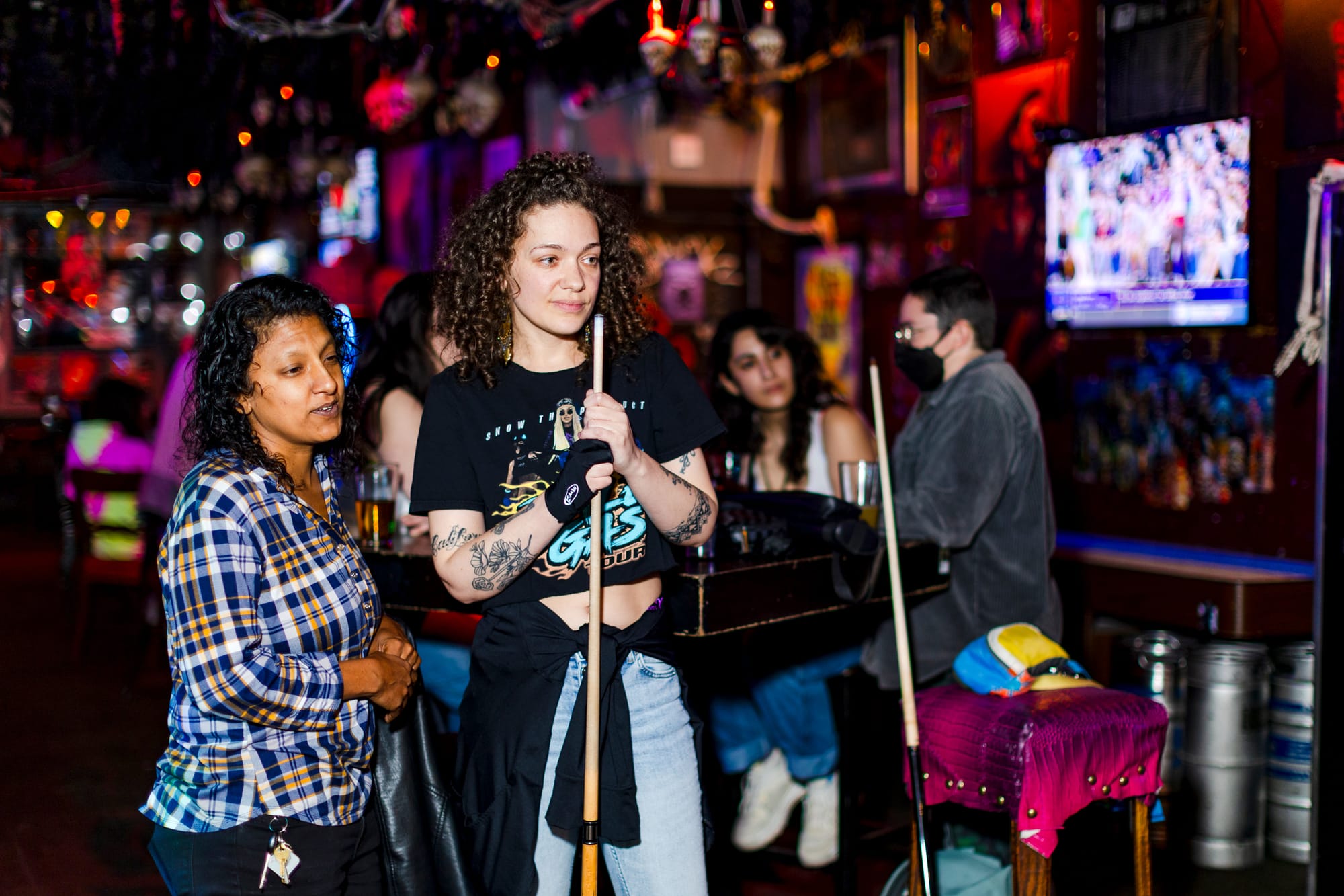 two people with long curly hair stand with pool cues waiting to play at a bar