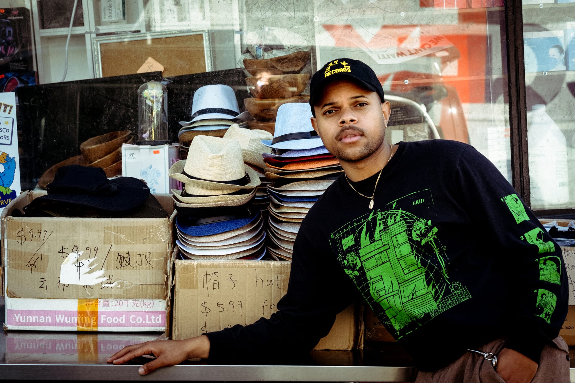 a man in a black and green shirt poses in front of a stack of hats on cardboard boxes