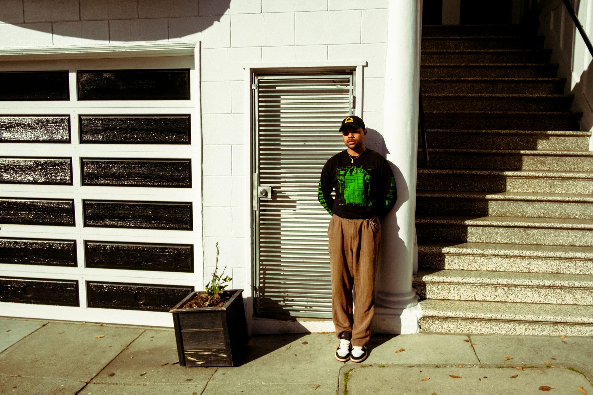 a young Black man in beige pants and a black and green shirt and black cap stands leaning agains a garage door on the street in San Francisco