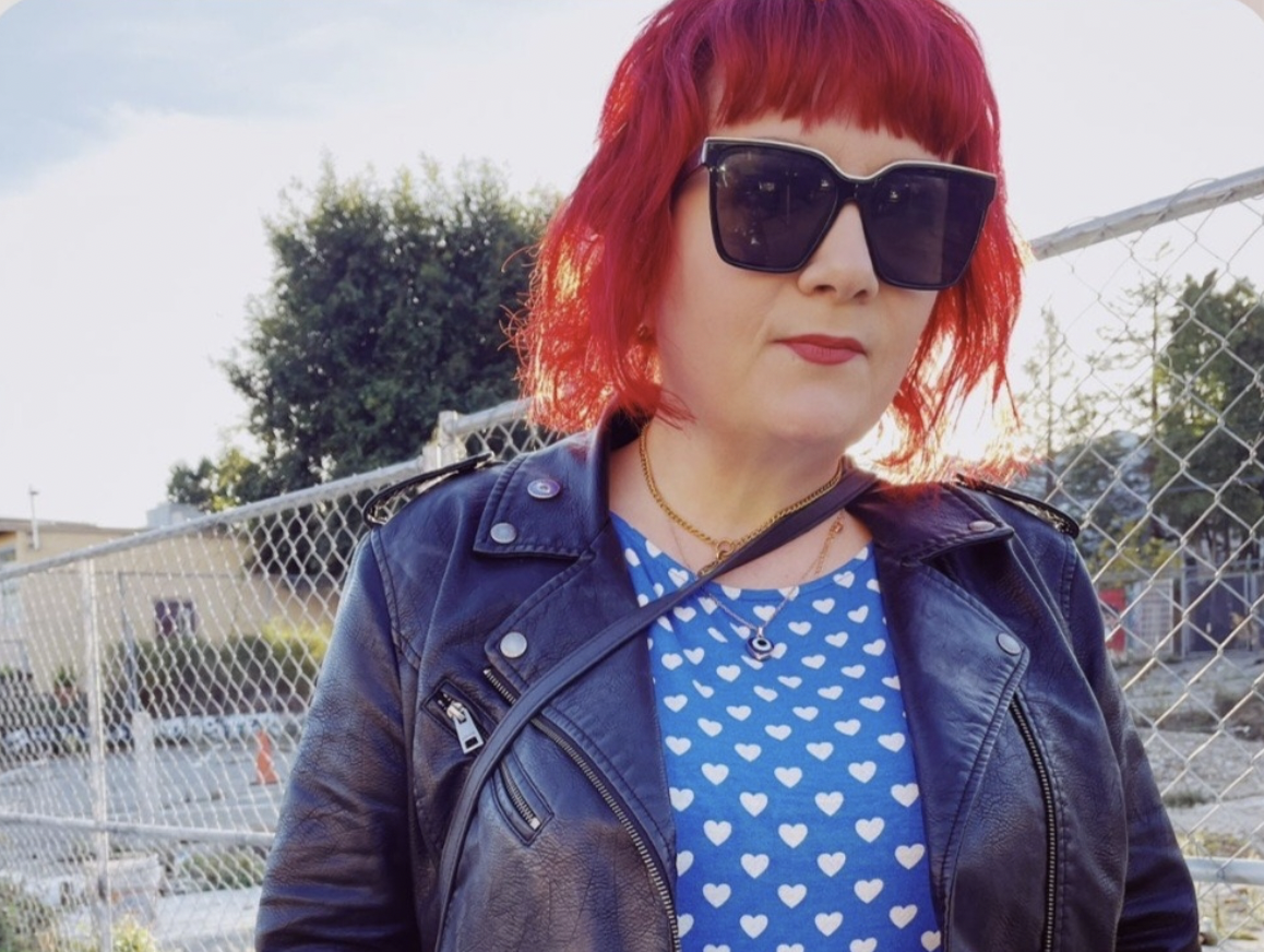 a woman in a blue dress with white hearts and a leather jacket with pink hair and sunglasses stands outside in front of a chain link fence