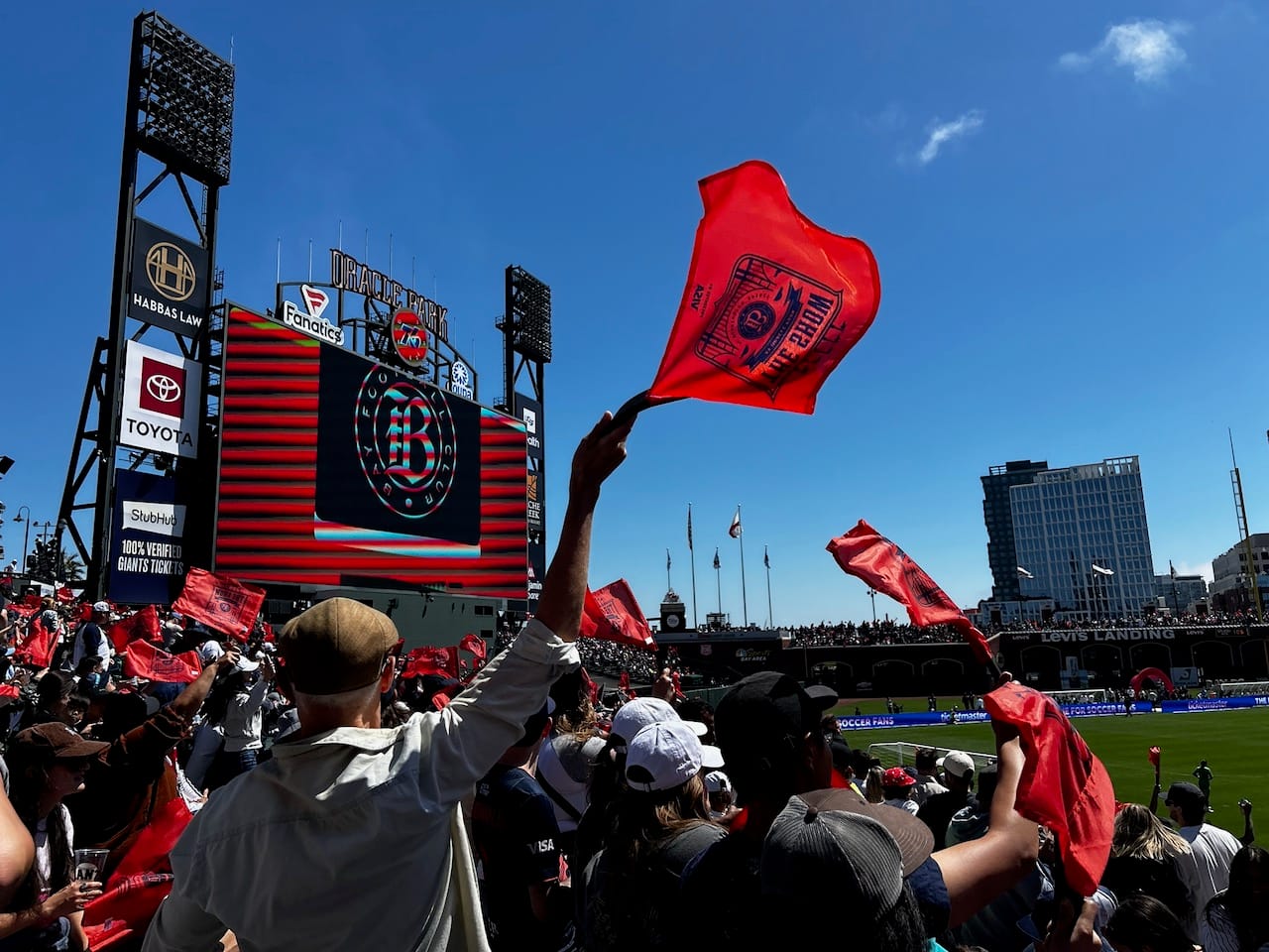 A crowd waving red, BayFC flags at Oracle Park on a sunny, nearly cloudless day.