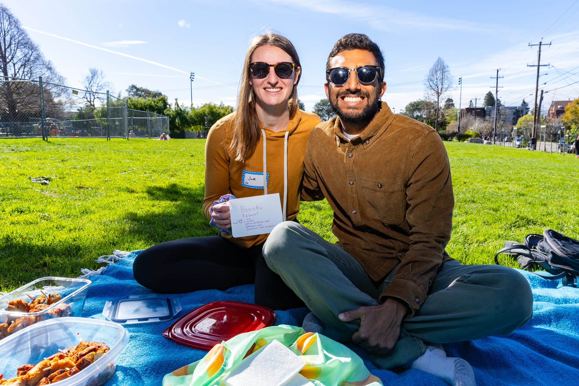 A man and a woman sit on a picnic blanket in a park surrounded by tupperware containers of food