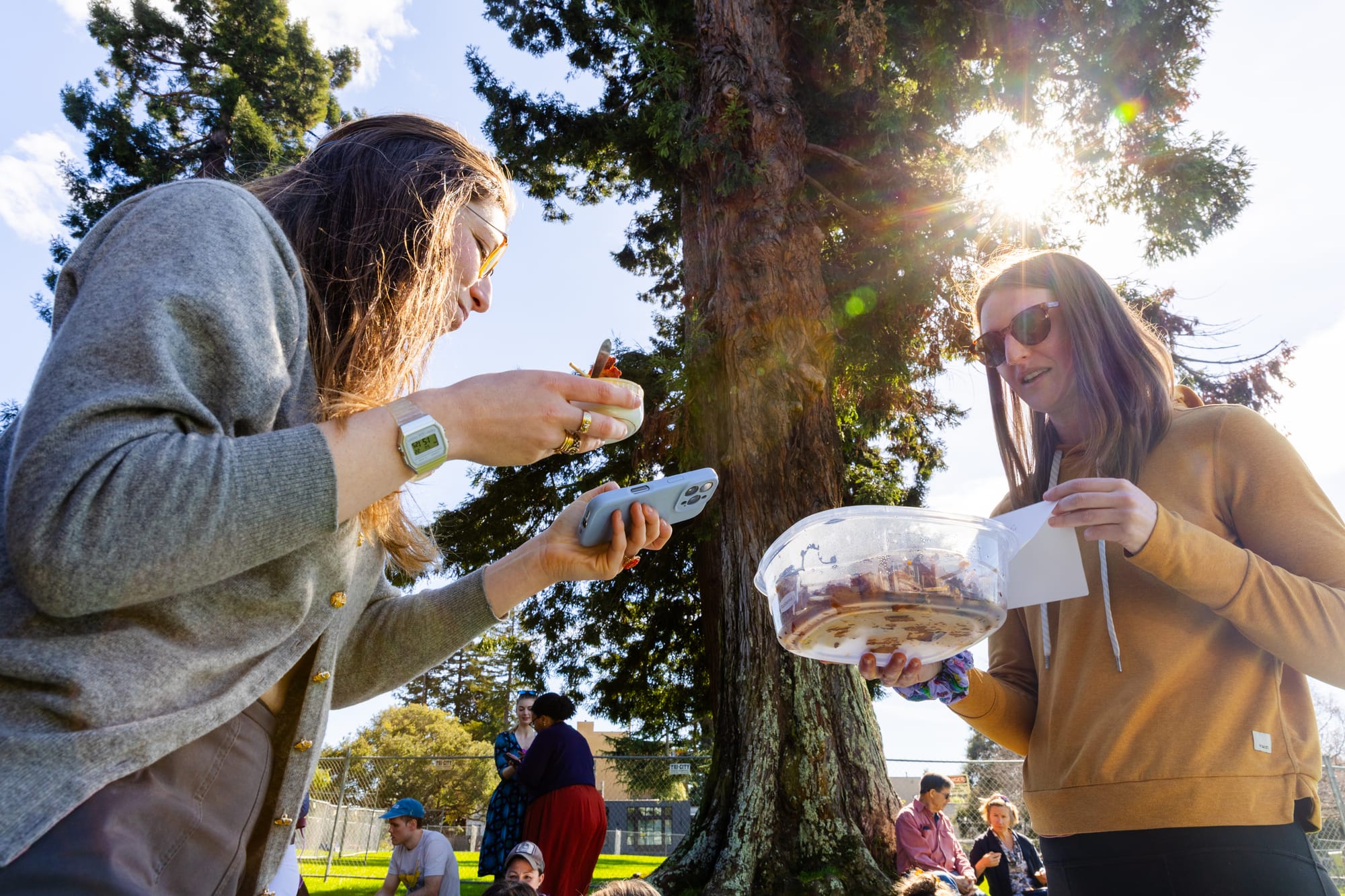 Two women stand under a tall tree sampling different dishes