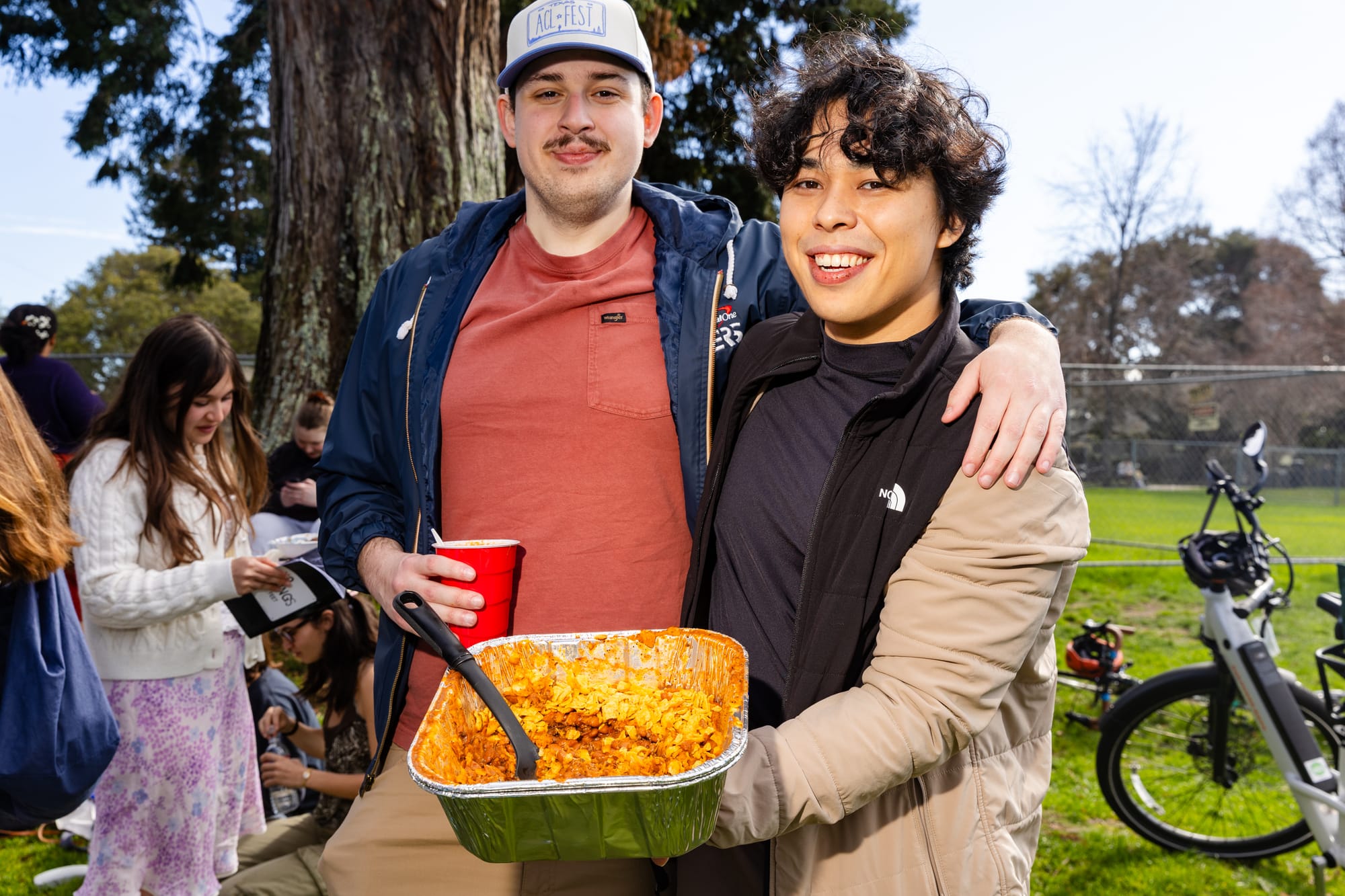 Two men stand with their arms around each other holding a container of frito pie
