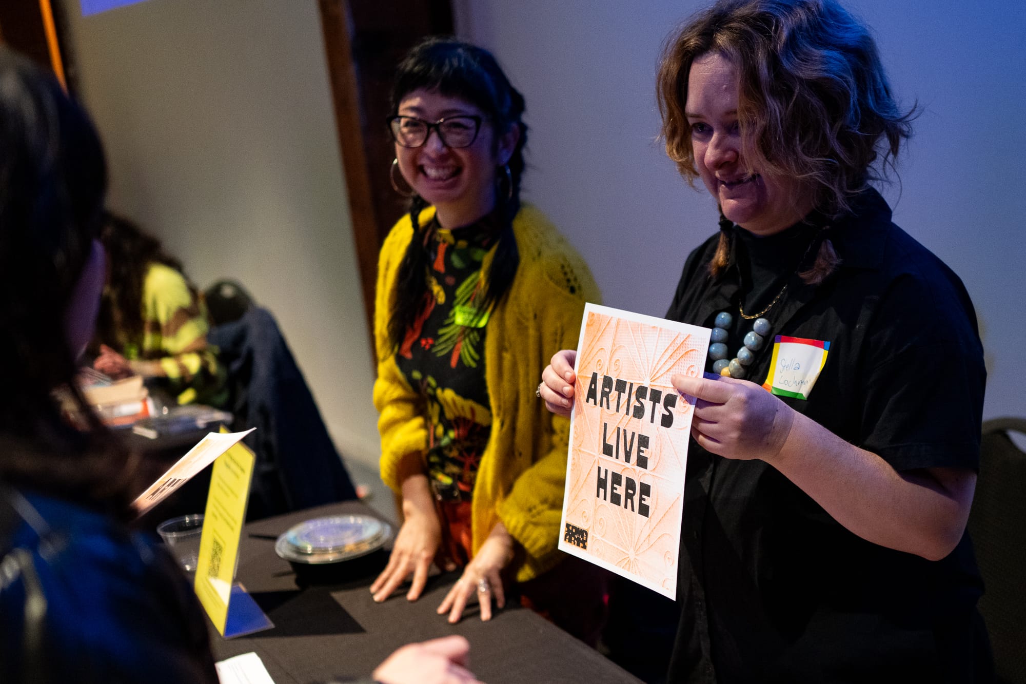 two people behind a table, one wearing a yellow sweater and braids, one wearing a black sweater and holding a sign that says 'artists live here'