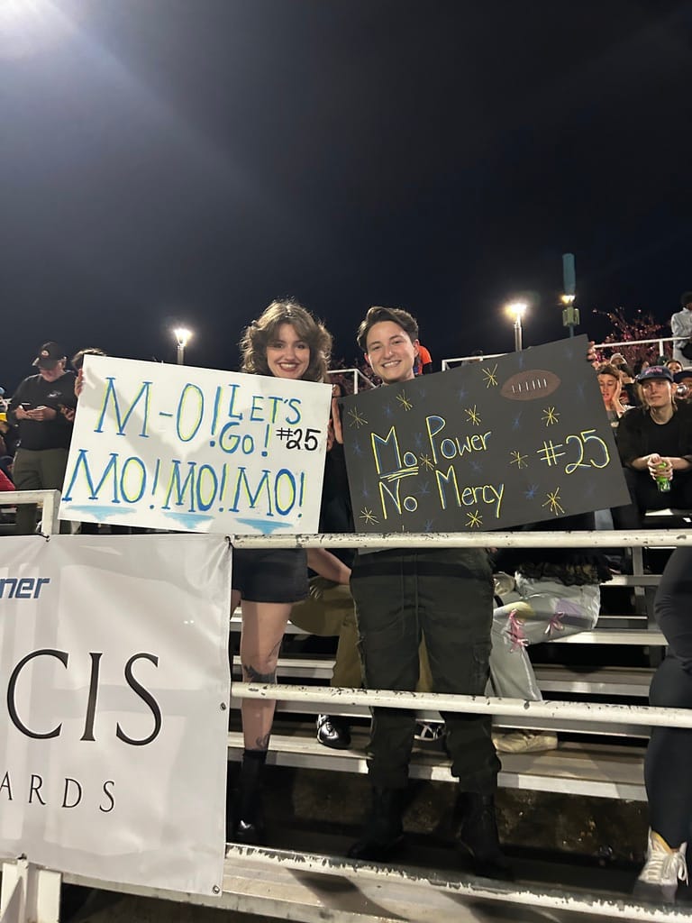 Two people smiling and holding signs that say "MO! Let's GO! #25 MO! MO! MO!" and "Mo Power No Mercy #25"