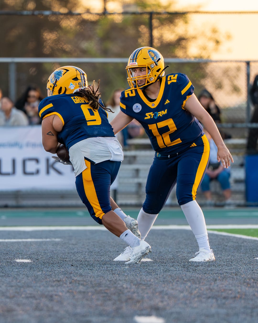 A quarterback hands the ball off to a running back, both are wearing blue uniforms with yellow helmets, and the running back has the ball secured and is running away.