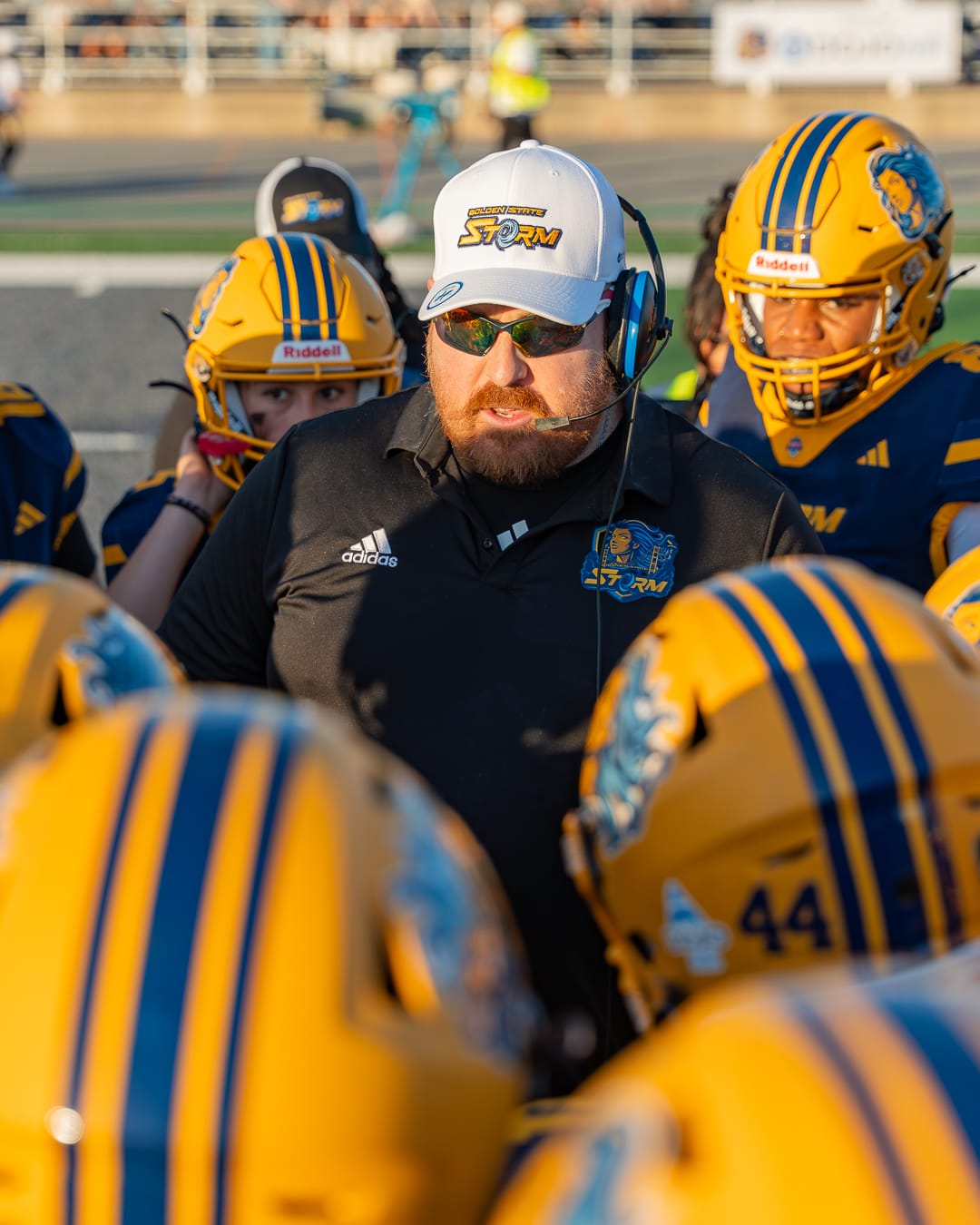 A man in a white baseball hat that says "Golden State Storm" wearing sunglasses and a headset, talks to the players around him.