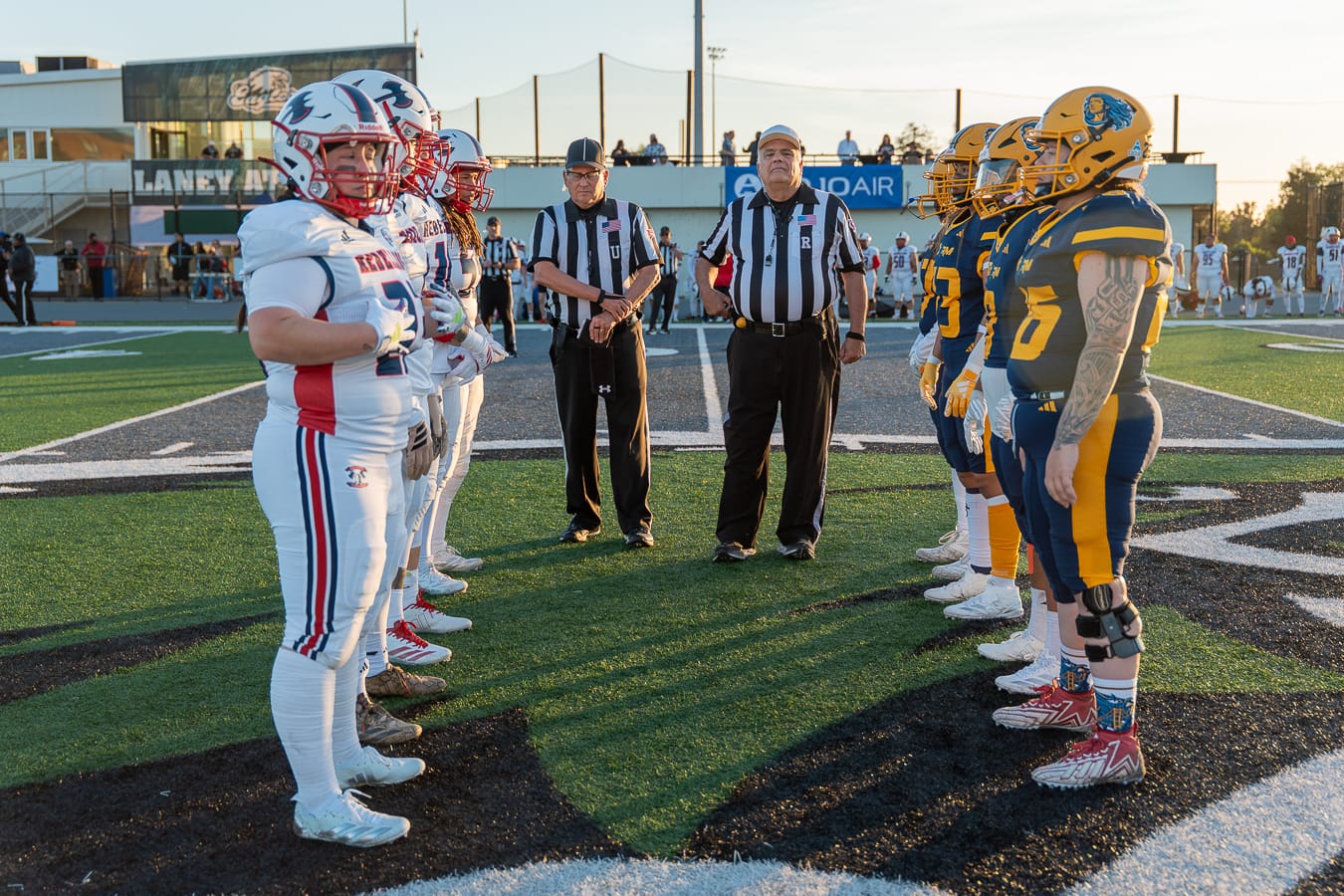 Two sets of football players line up facing one another, with referees in the middle. On the right, the team wears blue and gold. On the left, the team wears white and red.