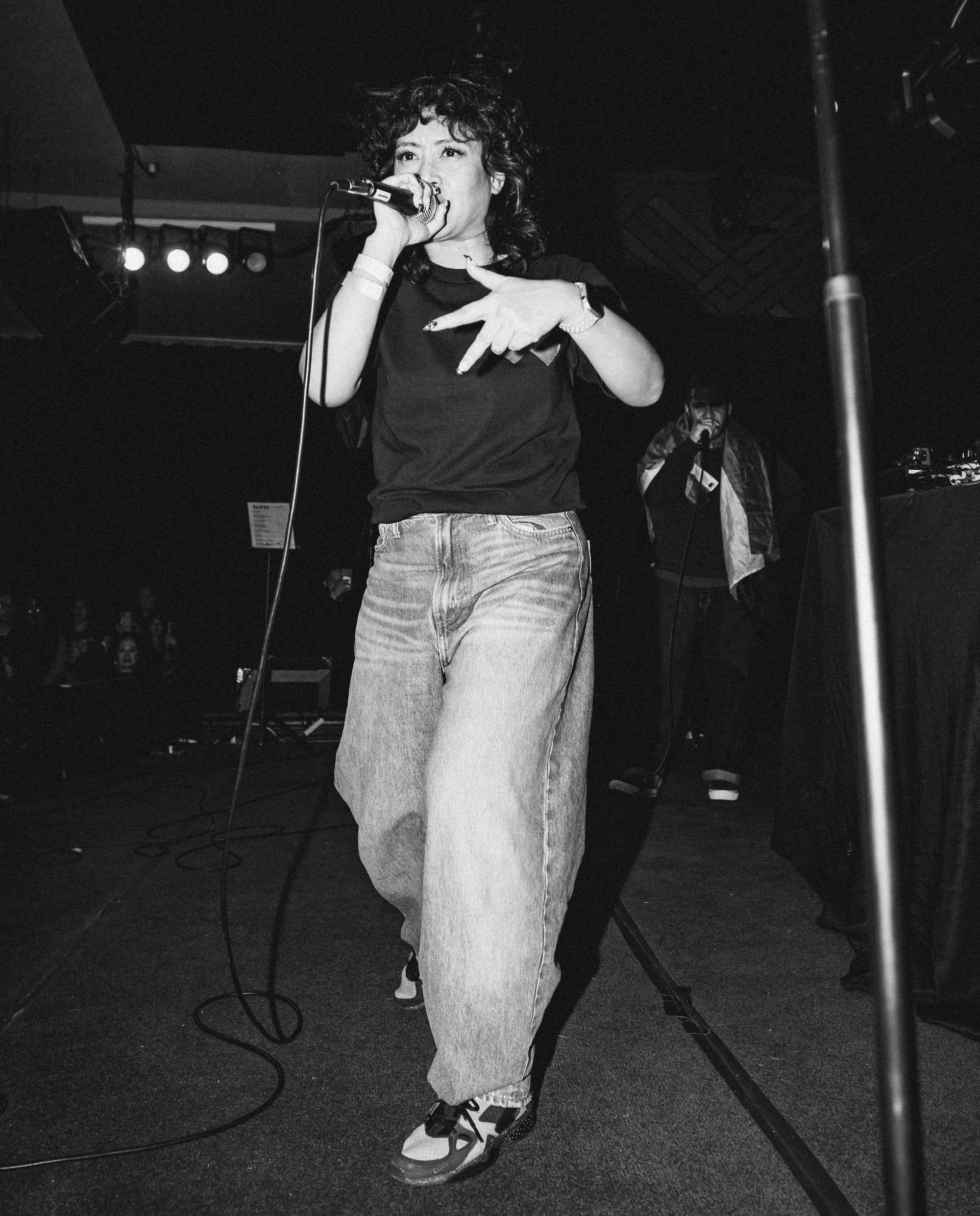 a black and white photo of a Filipina-American woman with a black shirt and baggy jeans and long painted nails, rapping into a microphone