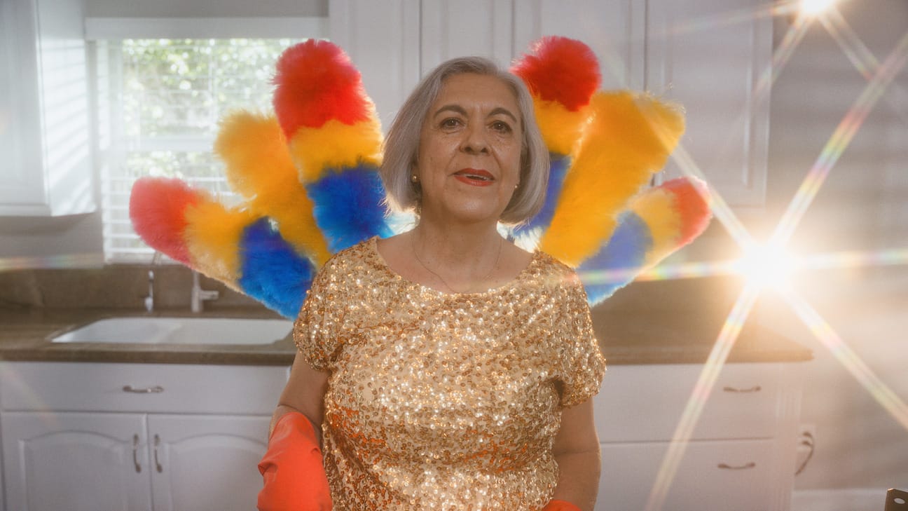 an older Latina woman in a sparkly gold shirt and orange gloves with brightly colored dusters fanned out behind her
