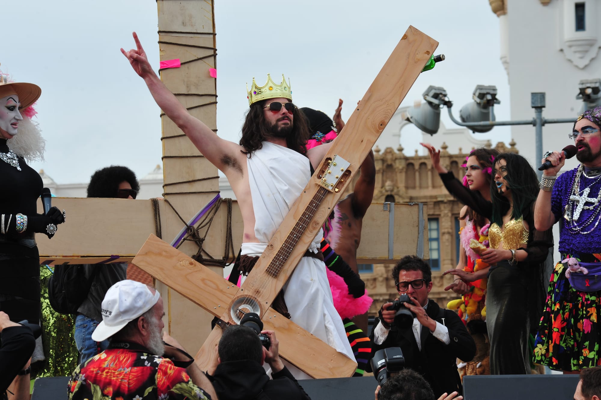a man dressed as Jesus with a guitar made of a giant cross wears a crown while drag performers and people cheer around him