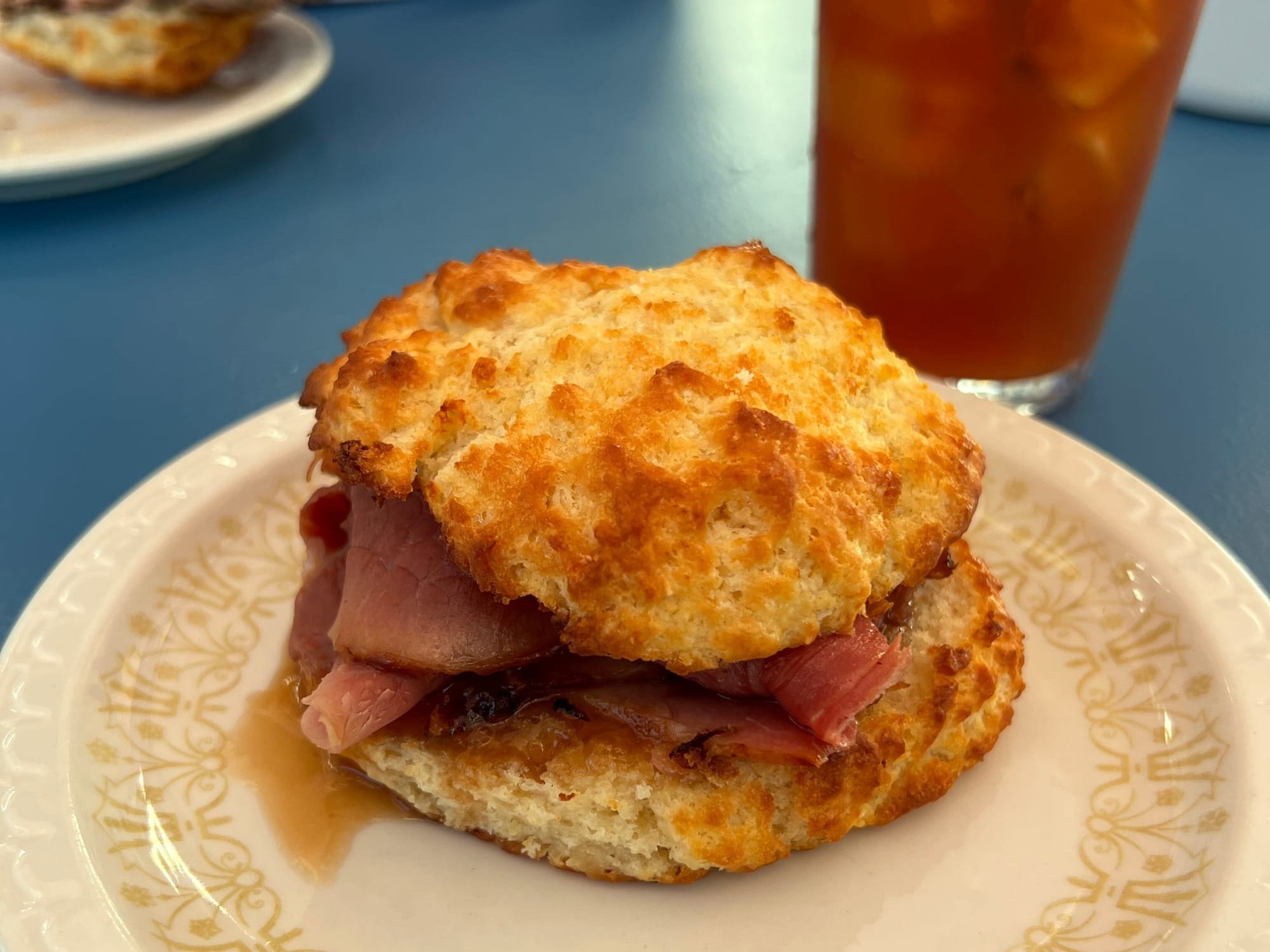 A craggy biscuit sandwich with ham on a white plate, with tea in the background.