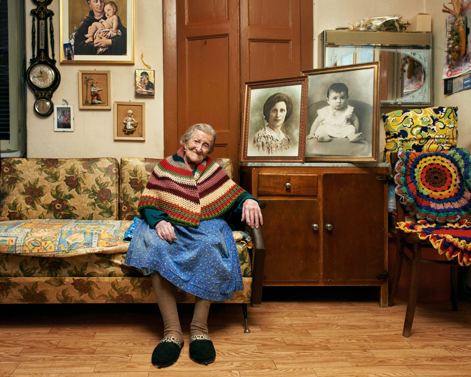 an old white woman sits on a colorful couch wearing a shawl, next to large black and white photos of family members