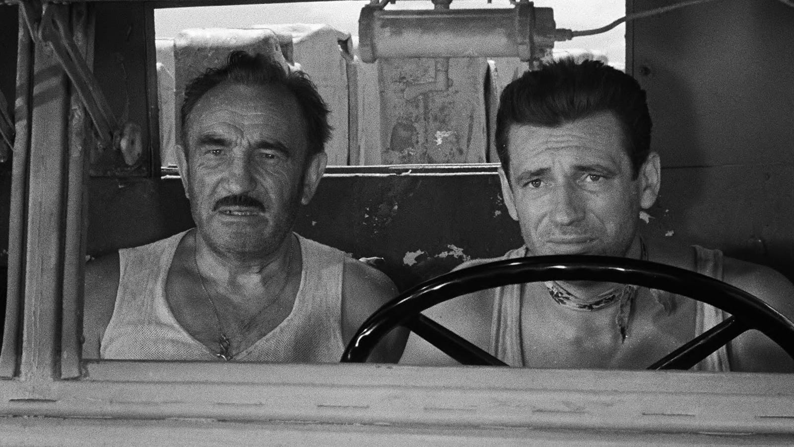 a black and white still image of two white men in a truck, one driving, both look worried