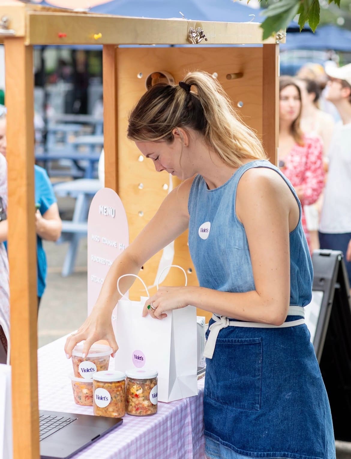 A blonde woman in a denim outfit stacks containers of beans on a table