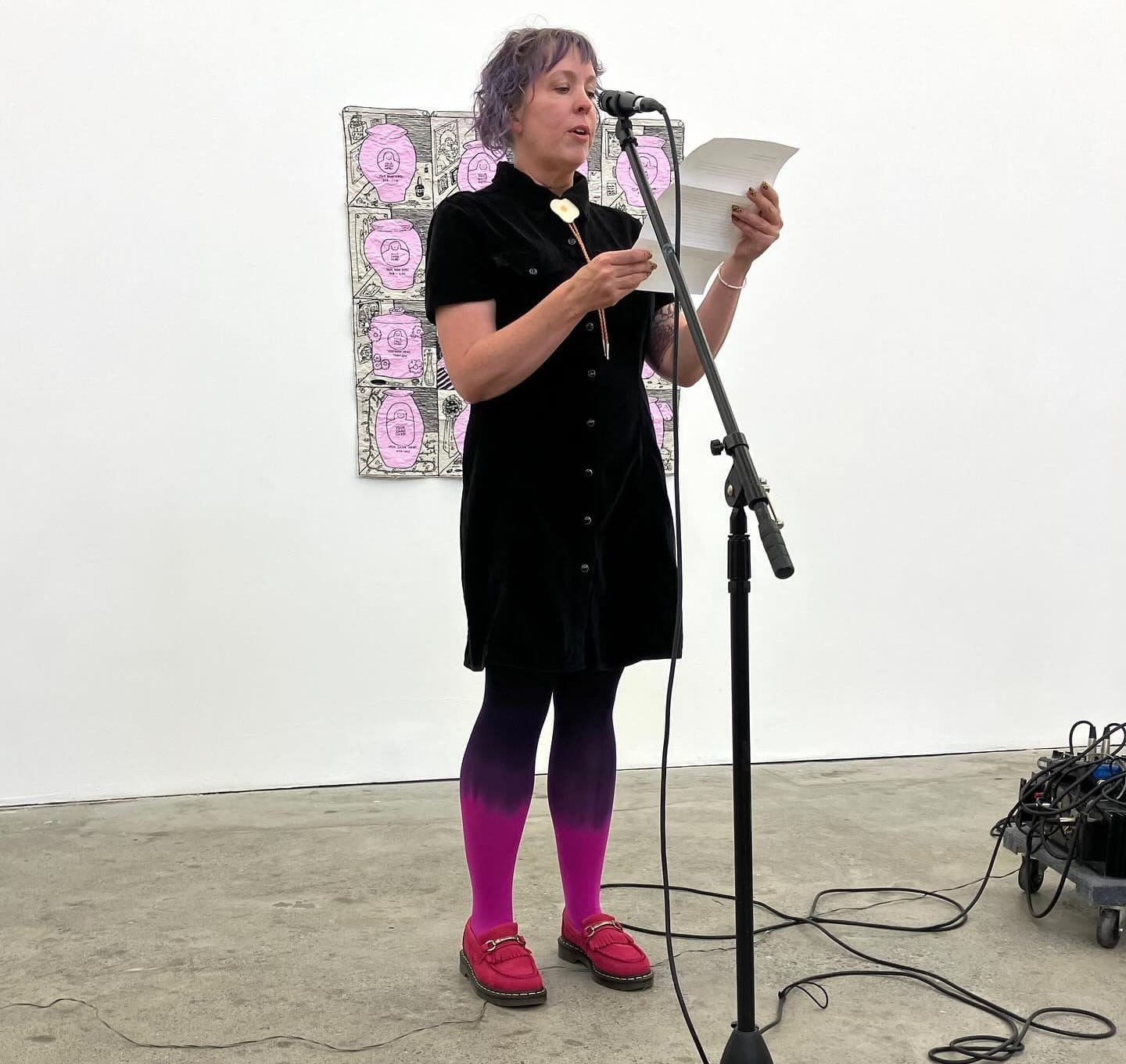 a woman with purple hair in a black dress and pink shoes reads a letter into a microphone