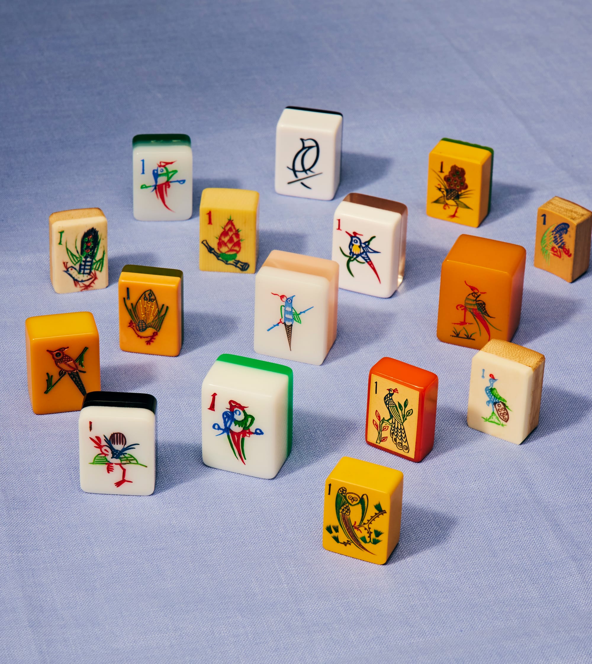 A collection of vintage and contemporary mahjong 1 bamboo tiles scattered across a light blue linen surface. Each tile depicts a different bird illustration — including peacocks, sparrows, herons, and parrots — in varied artistic styles. The tiles differ in material, age, and color, ranging from cream and ivory to amber, orange, and natural wood, reflecting sets from different eras and regions.