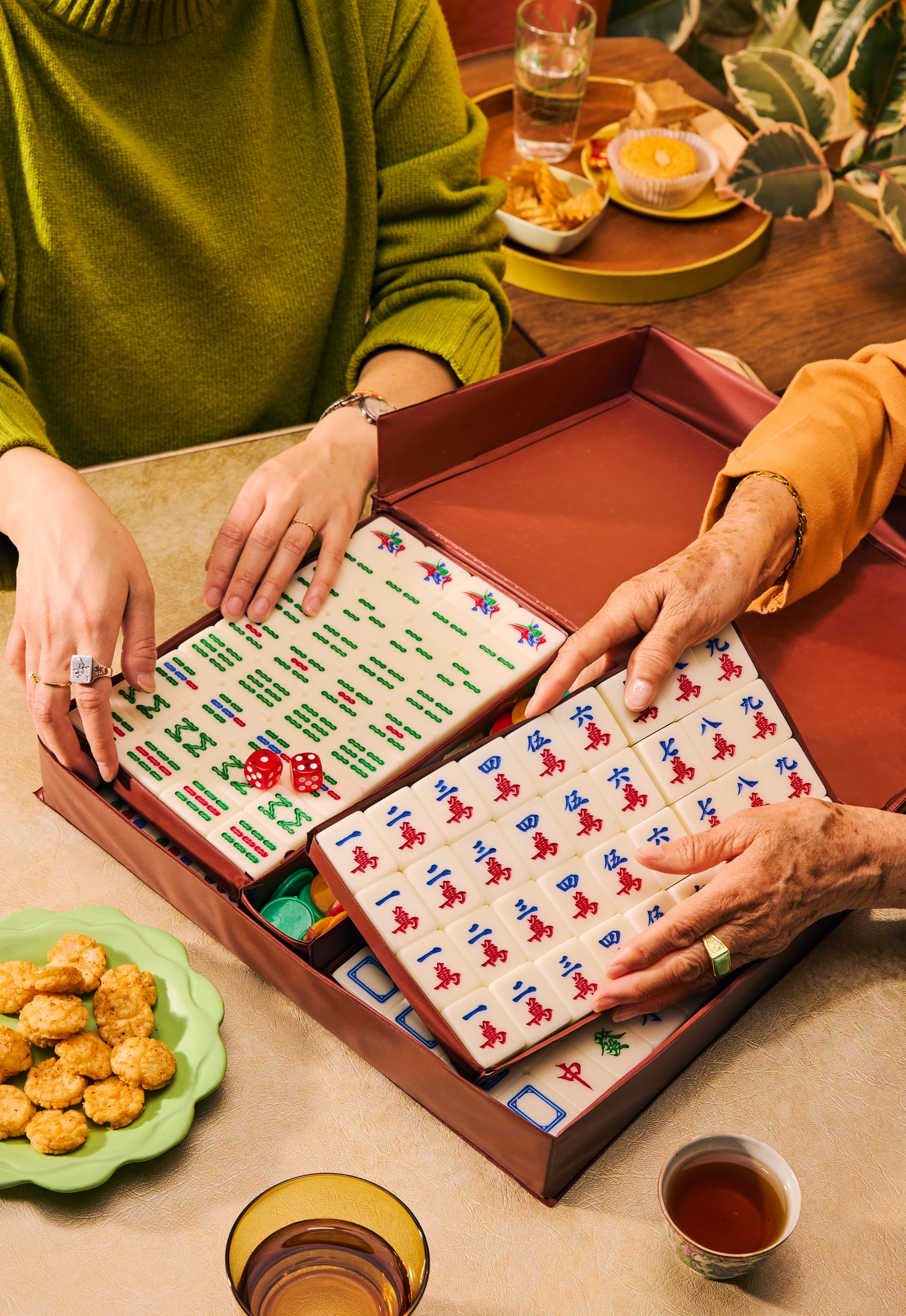 Two people open a red carrying case filled with mahjong tiles and red dice at a table. One person wears a green sweater and a ring, the other an orange sweater with an aged hand wearing a gold ring — suggesting an intergenerational gathering. Snacks, tea, a glass of water, and a candle are visible on the table nearby.