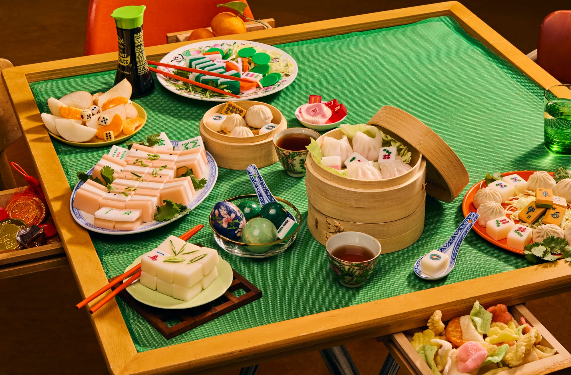 A wooden mahjong table with a green felt surface is covered with dim sum dishes and mahjong tiles arranged together as food styling. Items include a bamboo steamer filled with har gow dumplings topped with mahjong tiles, a plate of sliced pink terrine garnished with cilantro and embedded tiles, tofu-shaped tiles on a wooden rest with chopsticks, baozi buns in a smaller steamer, a dish of oranges and fruit with dice, a bowl of jade-colored eggs, and a plate of char siu-style items with tiles. Soy sauce, tea cups, a green glass, and gold coins in a side drawer complete the spread.
