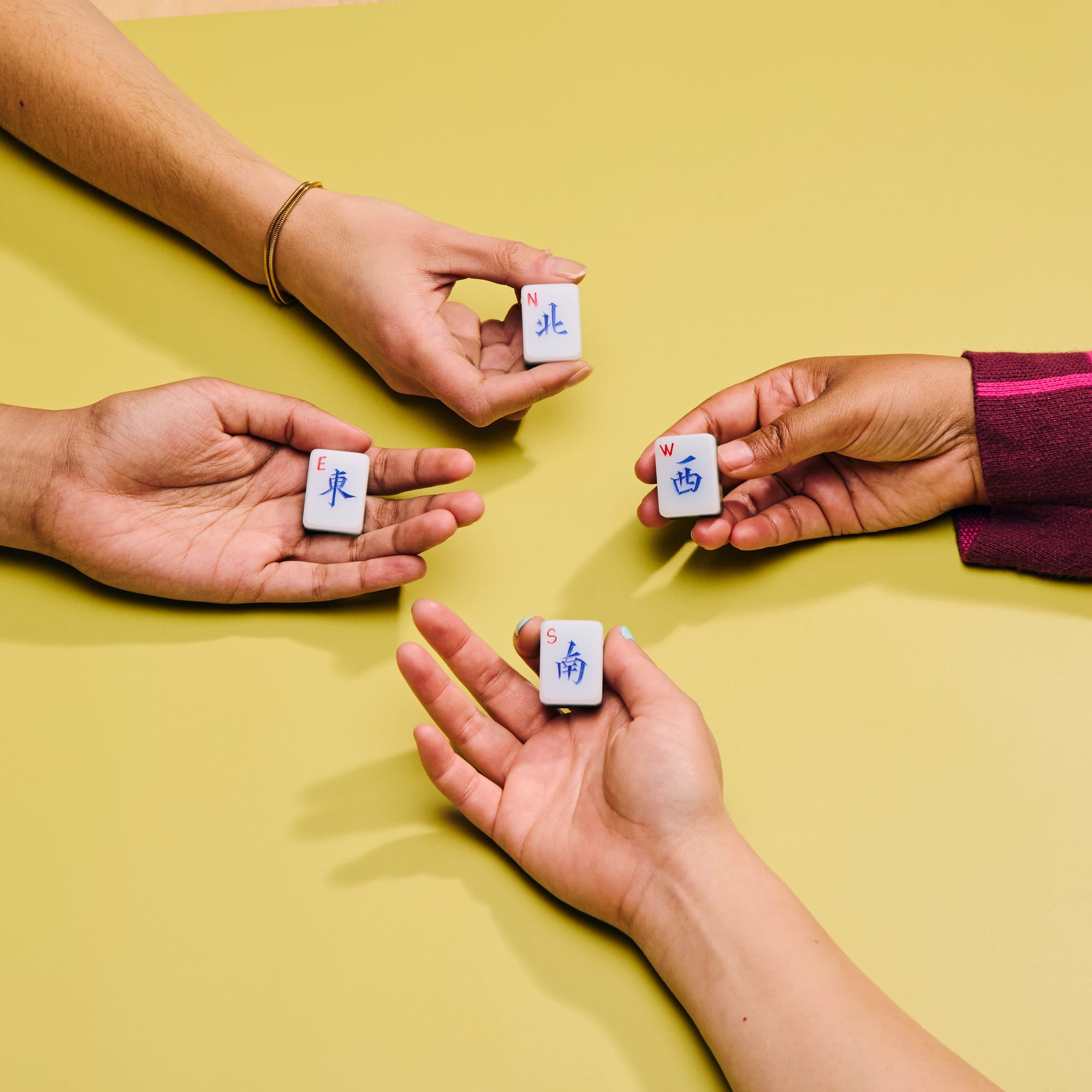 Four hands from different people reach toward the center of a yellow-green surface, each holding a mahjong wind tile: East (東), West (西), North (北), and South (南), labeled in both Chinese characters and English letters. The hands show diverse skin tones, and one wears a gold bracelet while another has blue nail polish and a third wears a burgundy knit sleeve.