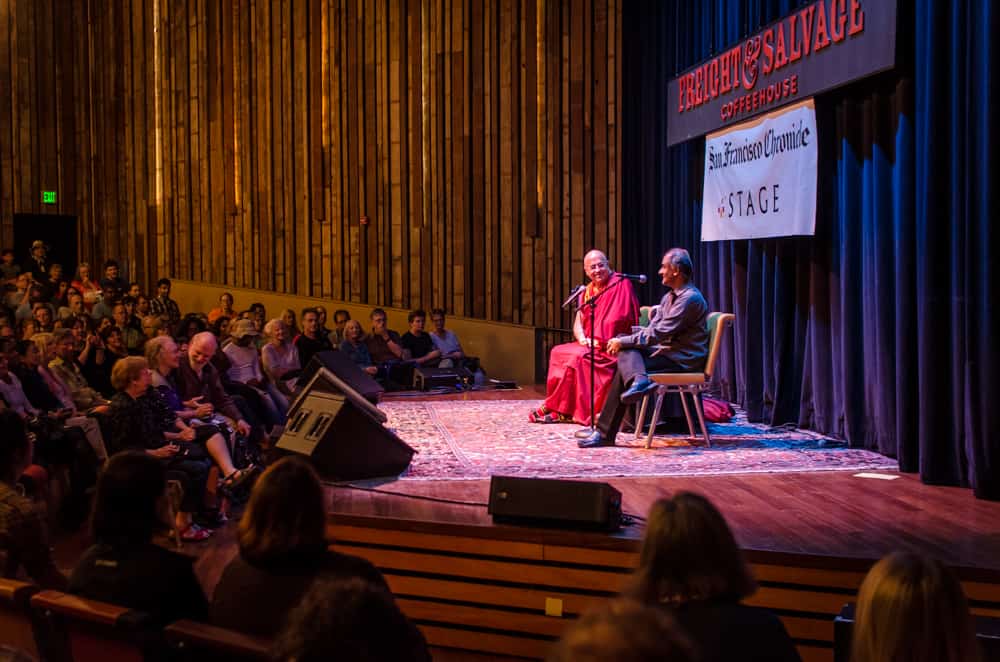 Two men — one a Buddhist monk in red and orange robes, one wearing a blue shirt — talk together while sitting on a stage in front of a crowd.