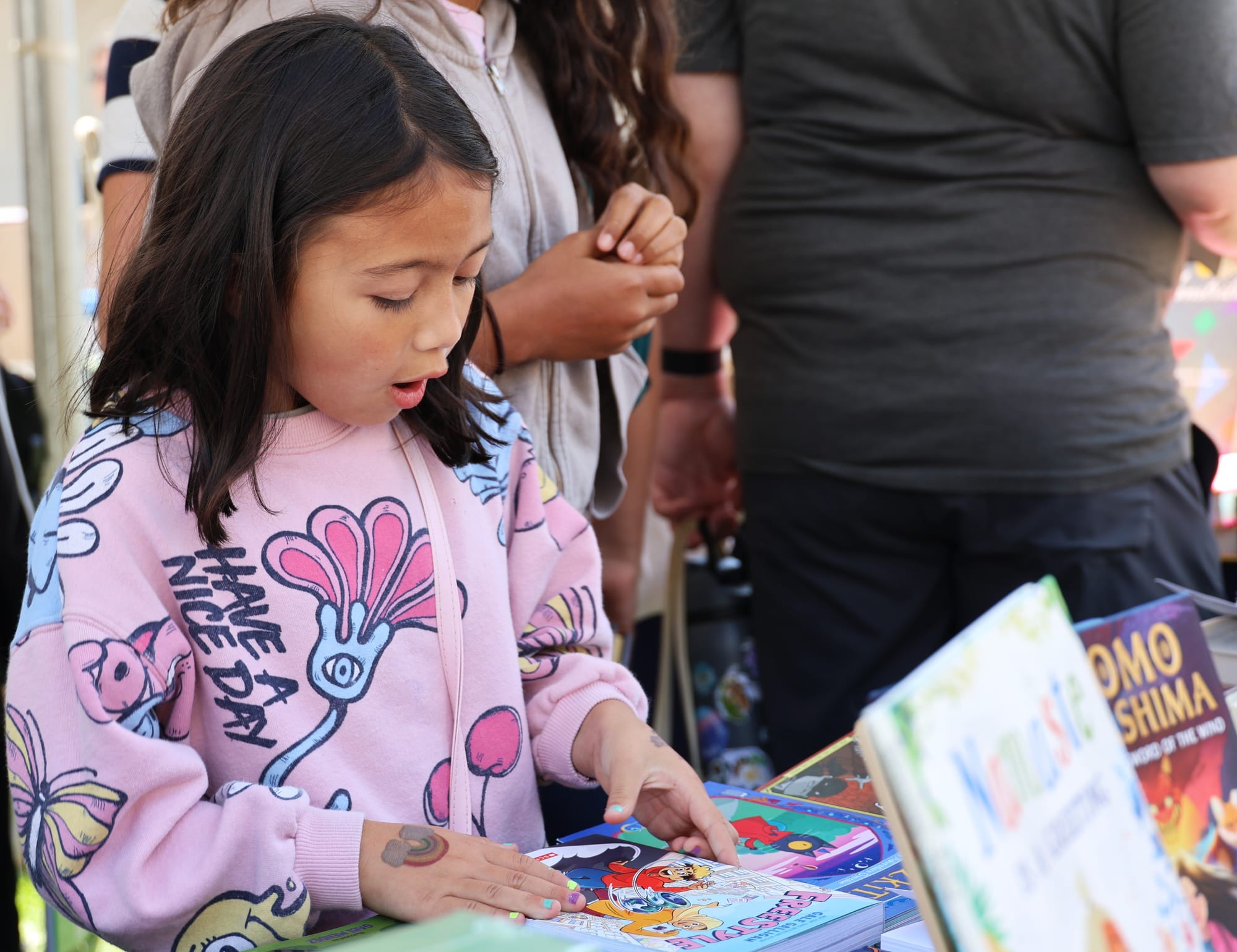 Child with long dark hair and a pink sweater that says "Have a nice day" touching a book at a book vending stall.