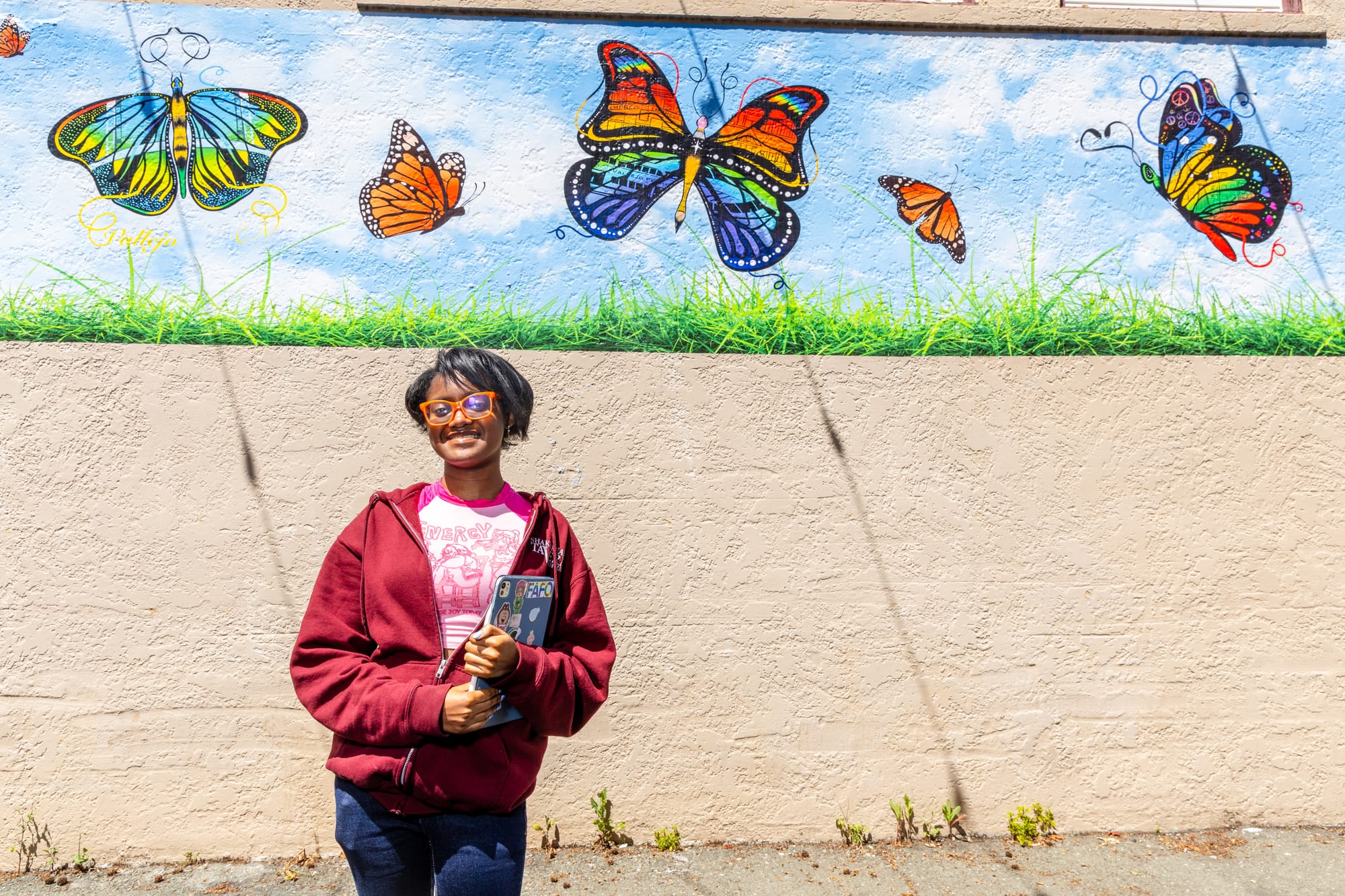 A young Black girl standing in front of a mural of butterflies, wearing a red sweatshirt, orange glasses, and holding an iPad.