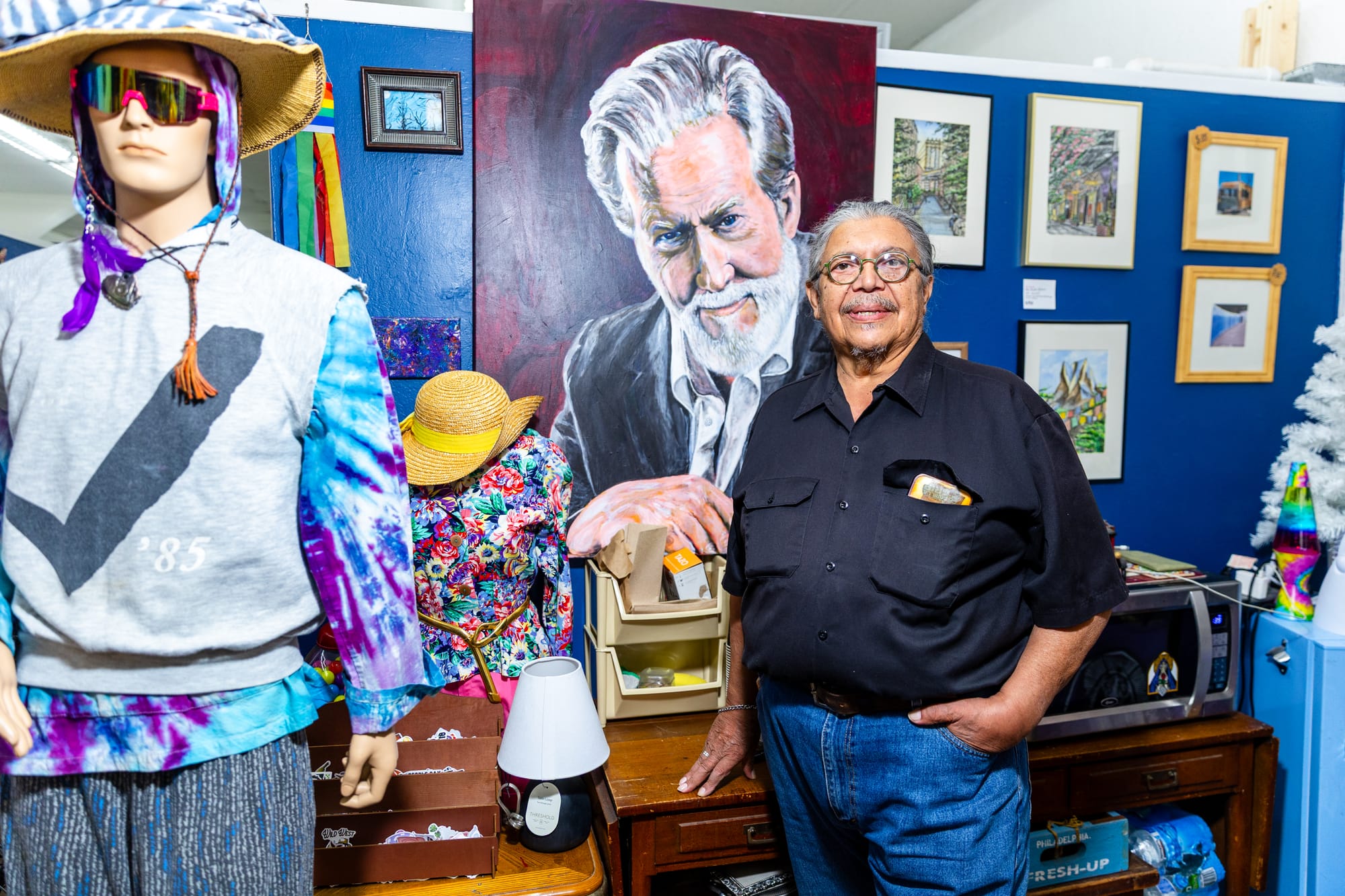 A man in a black shirt stands in front of a wall covered in art, including a large portrait he painted.