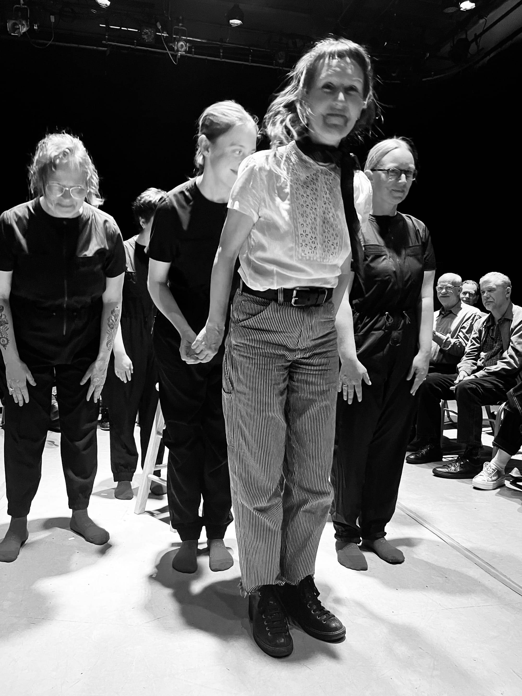 a black and white photo of a woman in striped pants smiling standing in front of performers all dressed in black at a musical performance