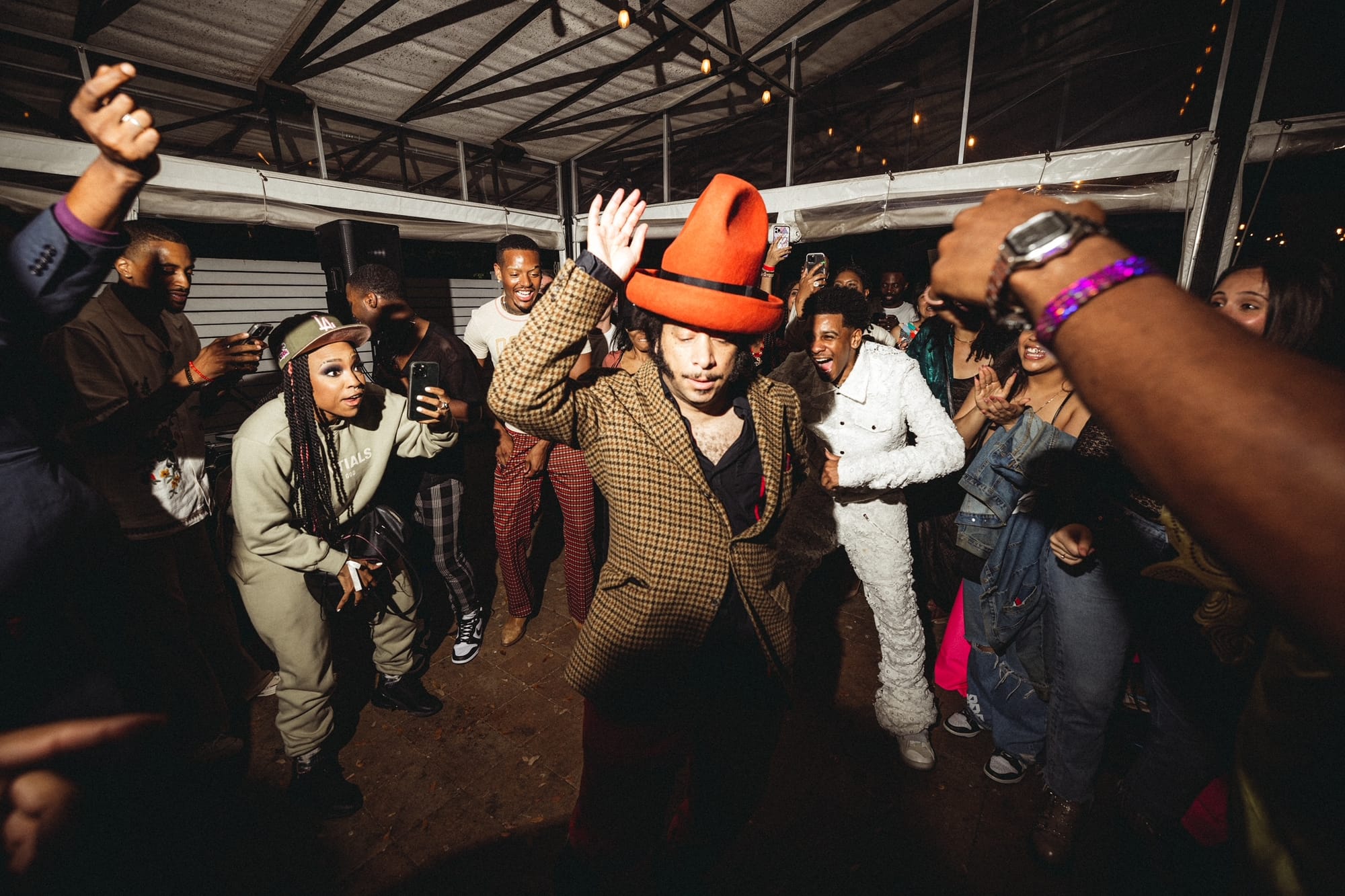Boots Riley, a man in a tall orange hat, dances in the middle of a circle while other people dance and cheer around him