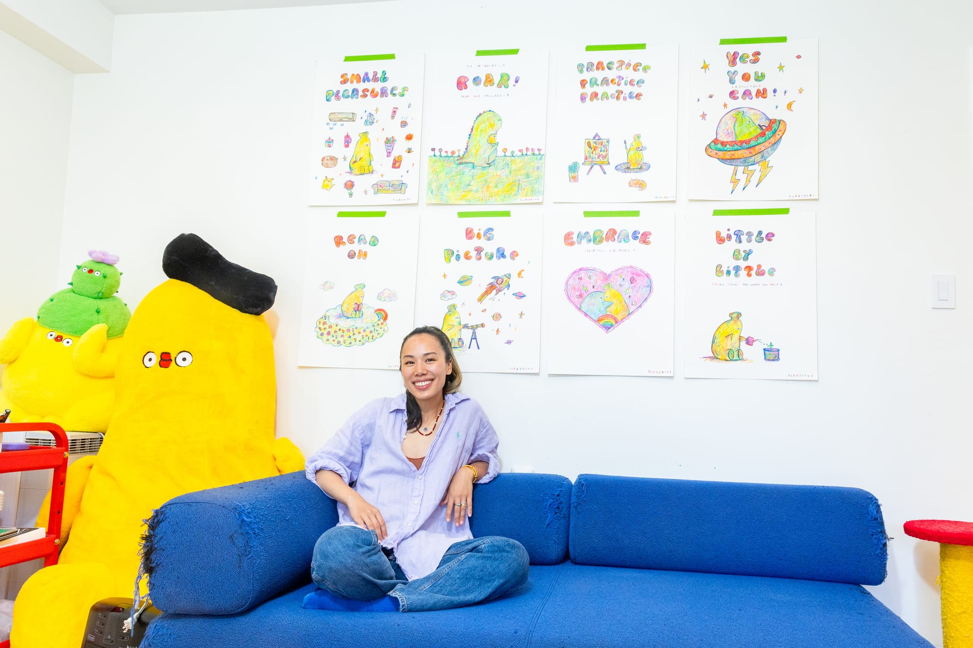 Woman with ponytail sitting cross-legged on a blue sofa in front of a wall of illustrations with text like "Small Pleasures," "Roar!" "Practice Practice Practice" "Yes You Can!" "Read On" "Big Picture" "Embrace" and "Little By Little"