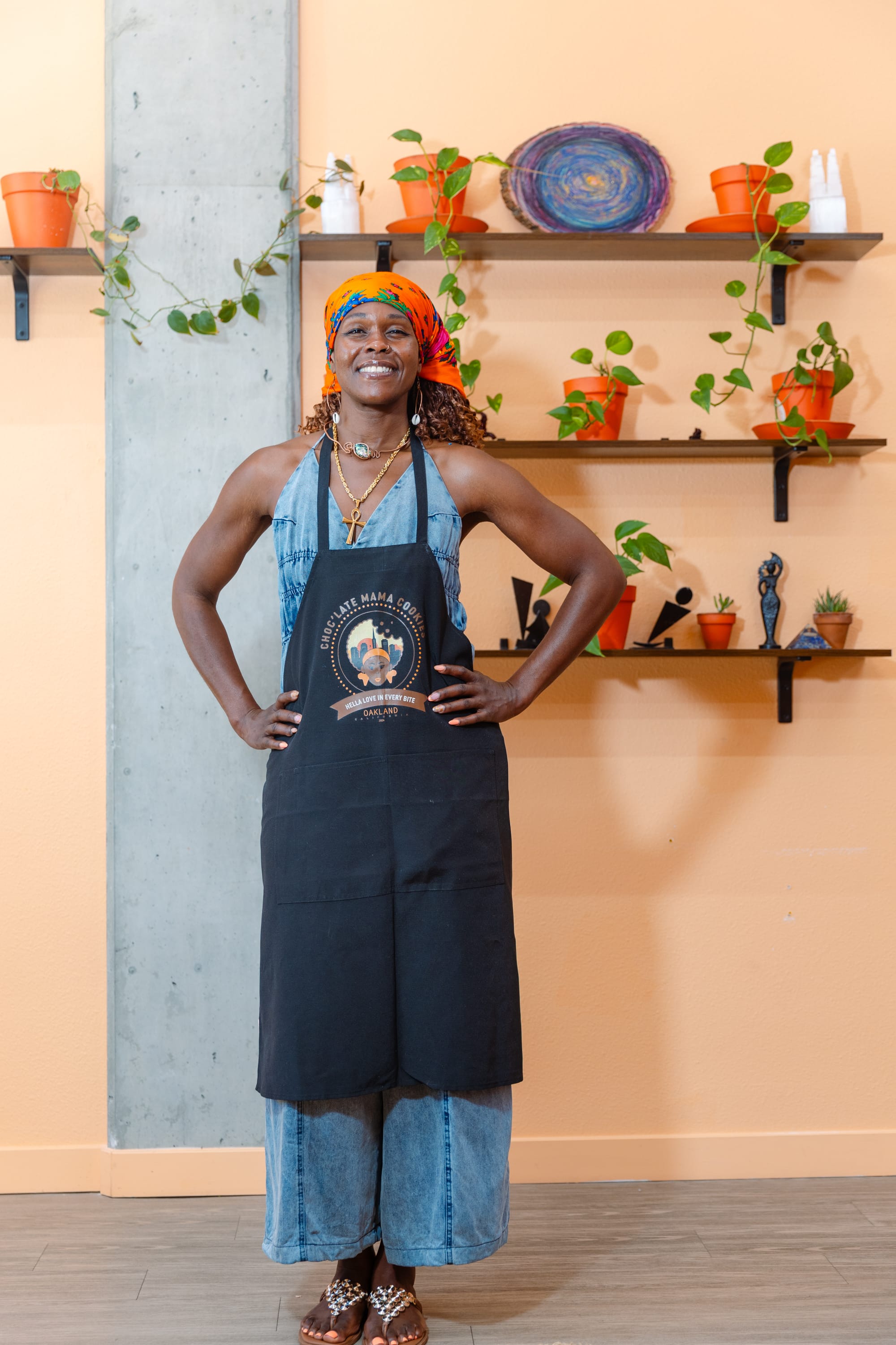 A woman wearing an orange kerchief on her head and a blue apron standing with her hands on her hips in front of shelves with statues and pothos plants.
