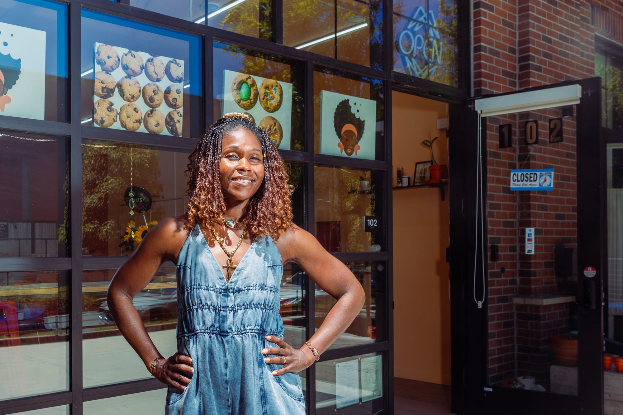 Woman in a denim one-piece outfit standing outside of a cookie shop with her hands on her hips.