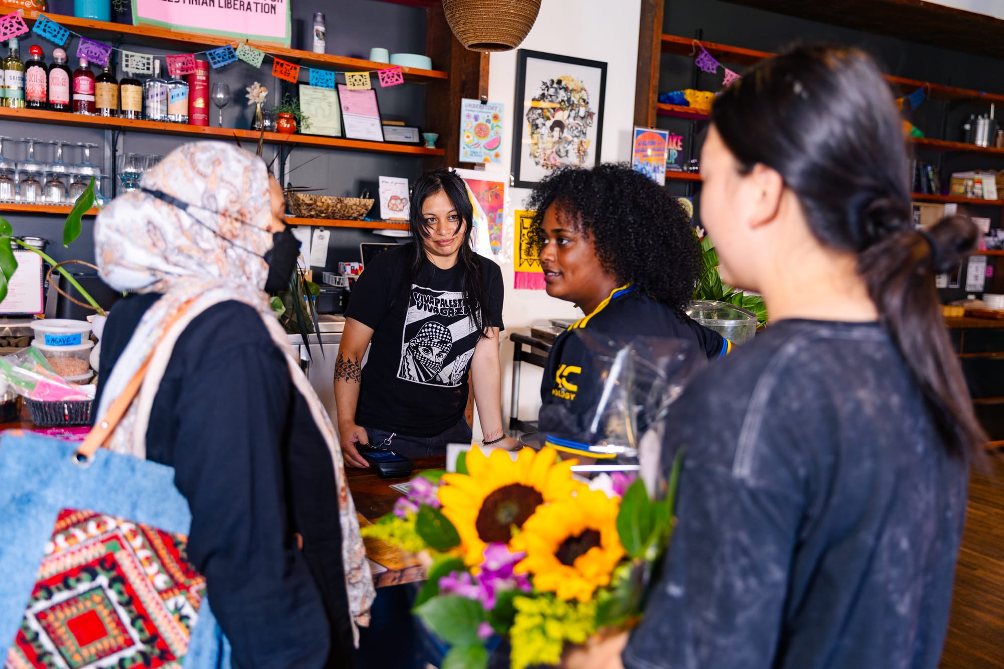 Person with long hair wearing a "Viva Palestina" t-shirt looks on while people stand at a restaurant counter.