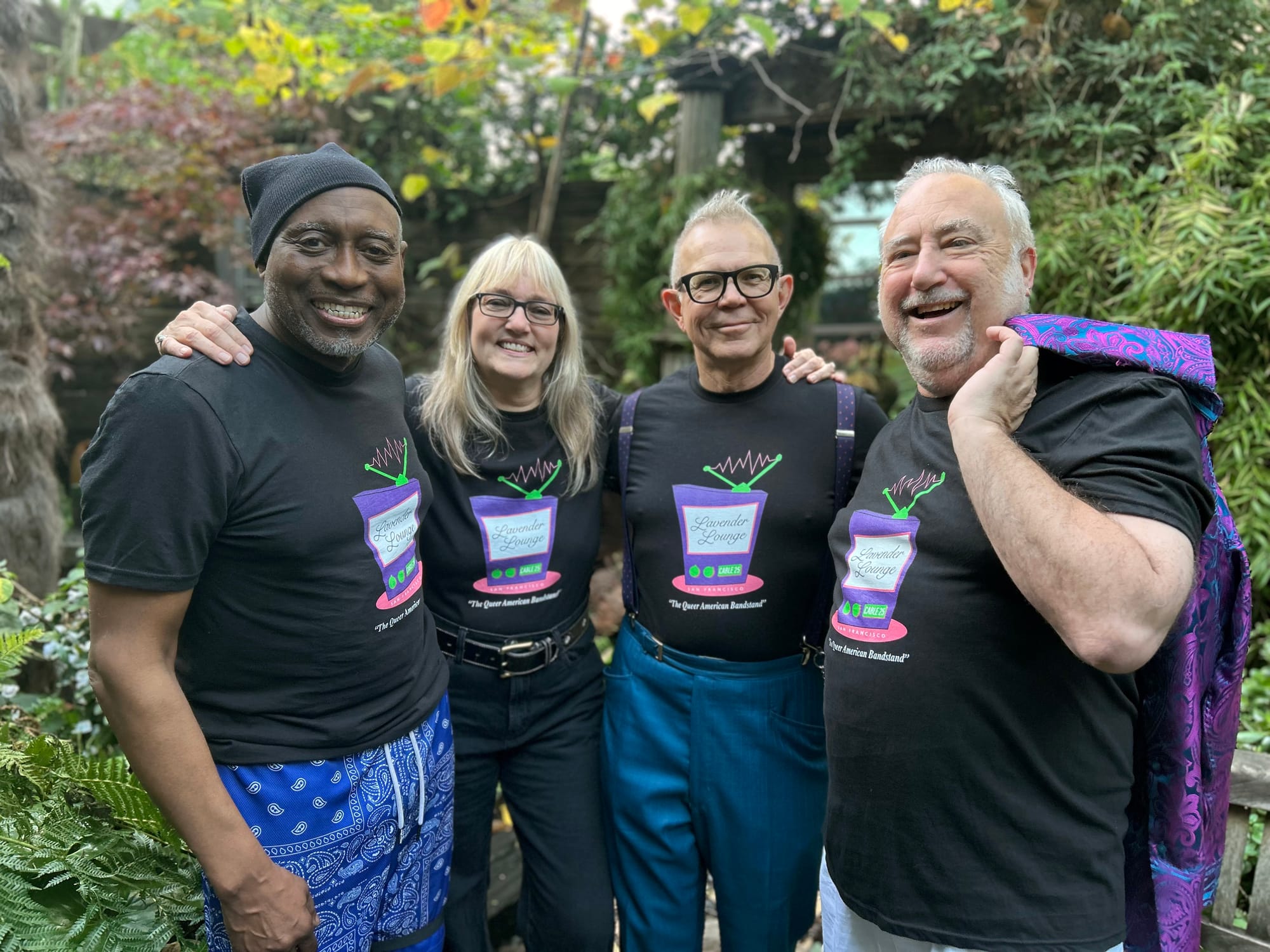 four people pose for a potrtait: a Black man in a beanie, a white woman with blond hair and glasses, a white man with glasses and suspenders, and a white man with a colorful cloth draped over his left shoulder. All wear T-shirts that read 'Lavender Lounge, the queer American Bandstand'