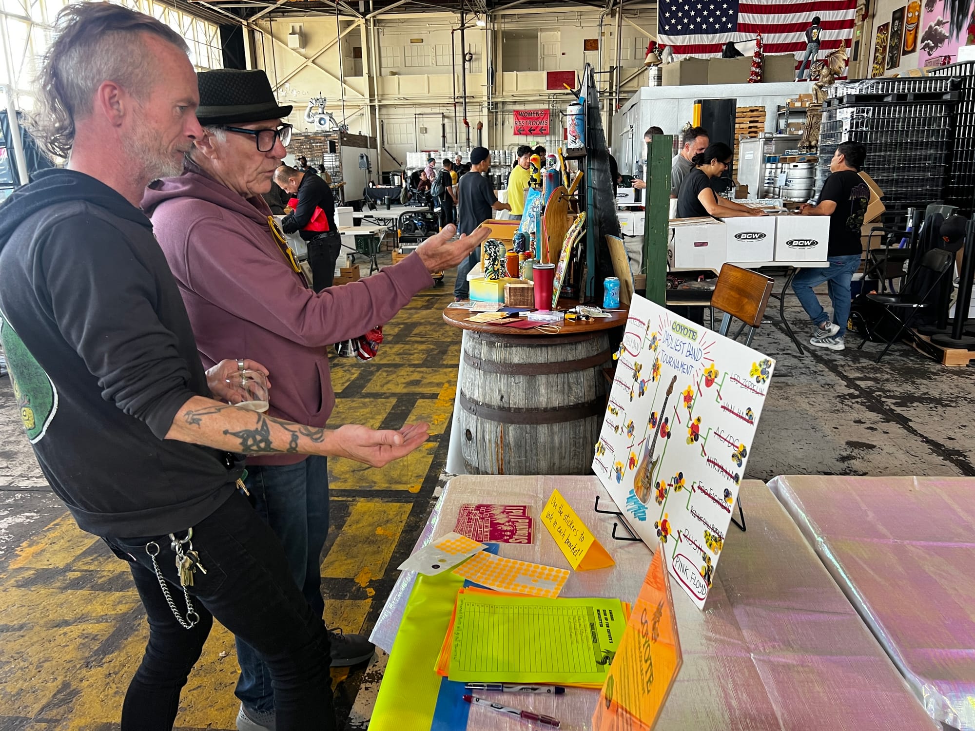 two men gesture at a bracket of dad bands at a record swap at a beer hall