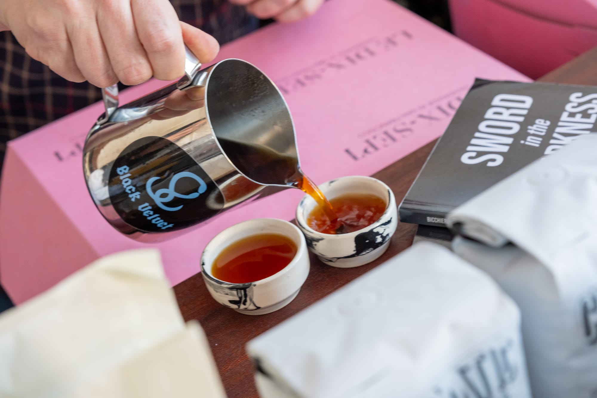 A person pours coffee out of a metal vessel into two small ceramic cups.