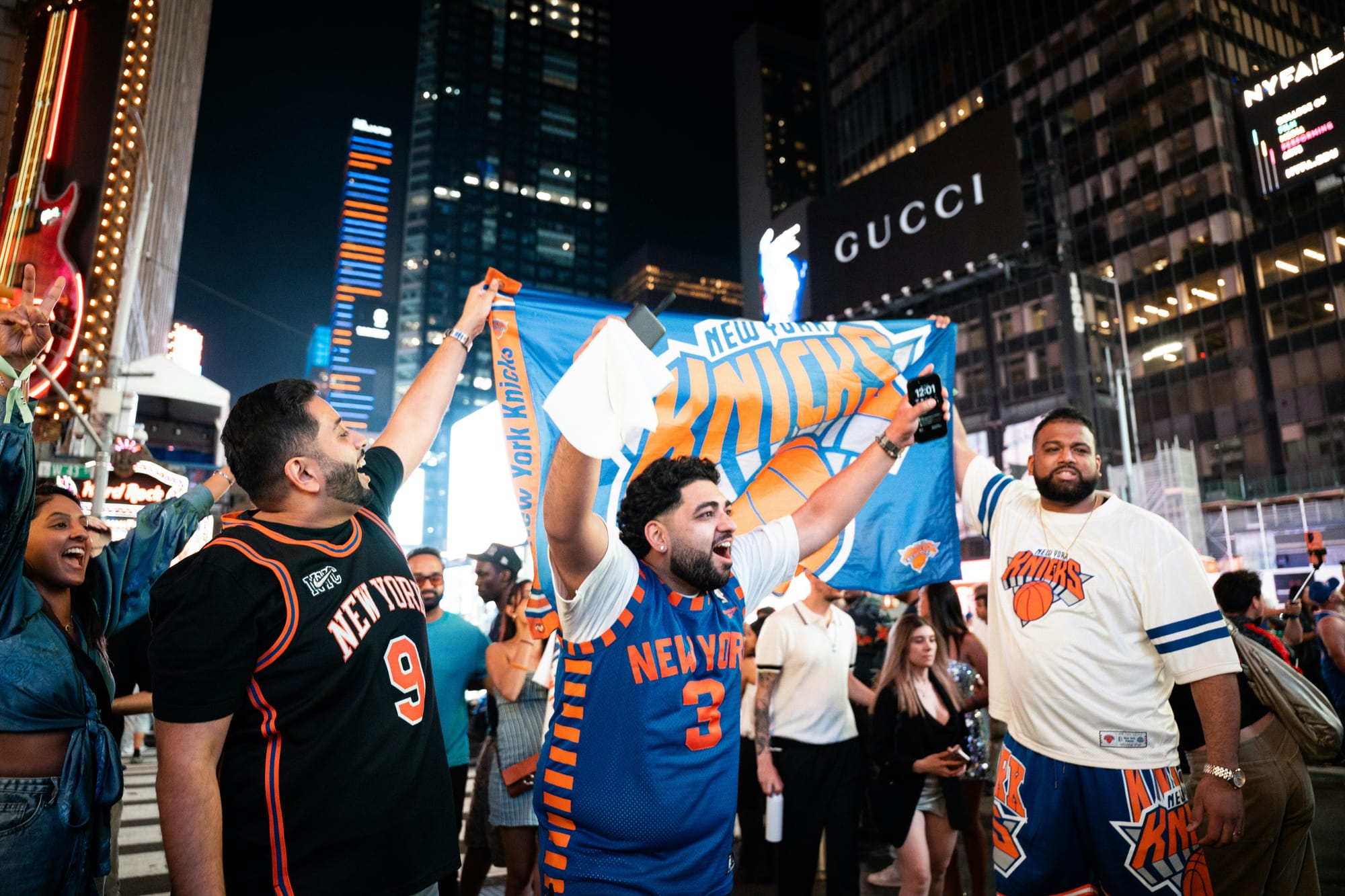 Three men in Knicks jerseys cheer while waving a Knicks flag in Times Square.