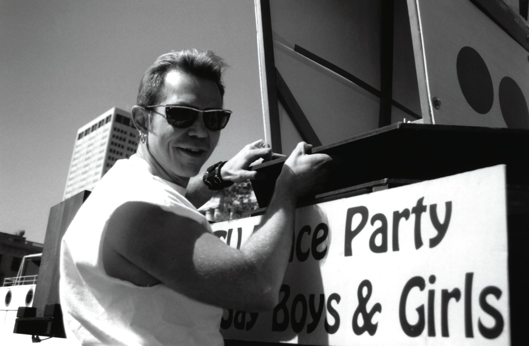 a black and white photo of a man in sunglasses hanging on the side of a float for a pride parade