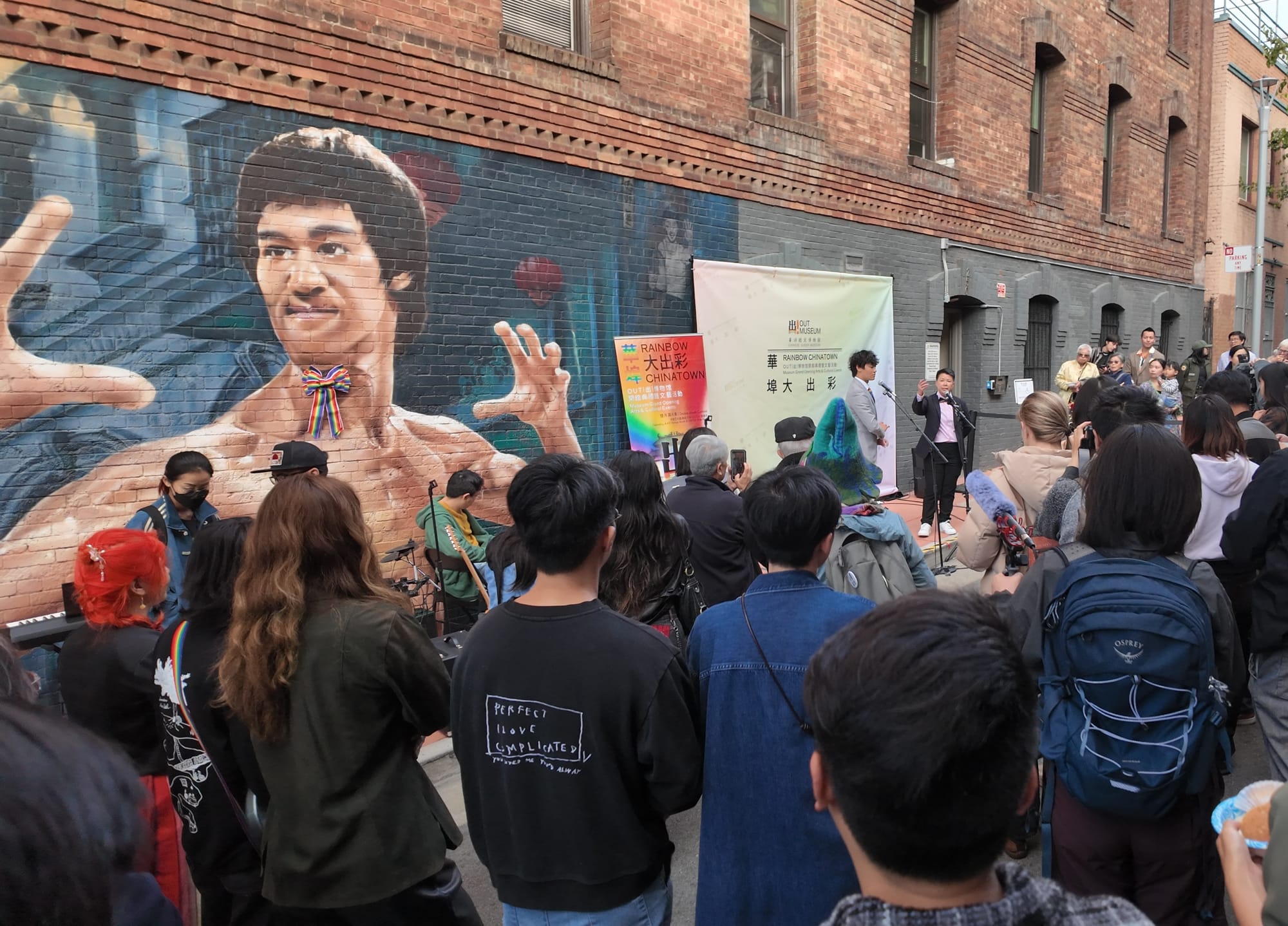 A crowd gathers in front of a Bruce Lee mural and watches two speakers at a mic to celebrate the opening of OUT Museum.