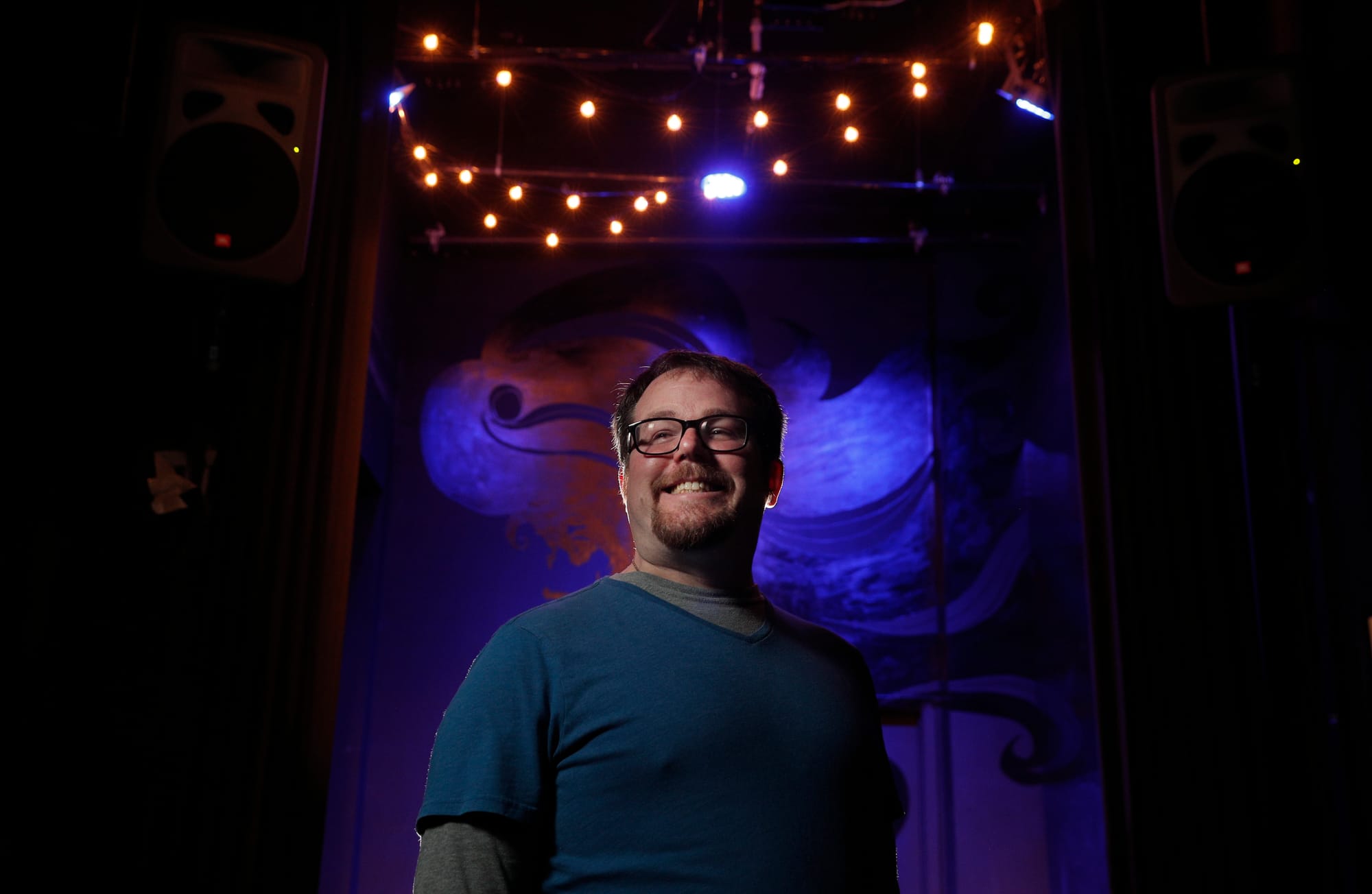 a white man in a blue shirt and glasses smiles in a darkly lit theater