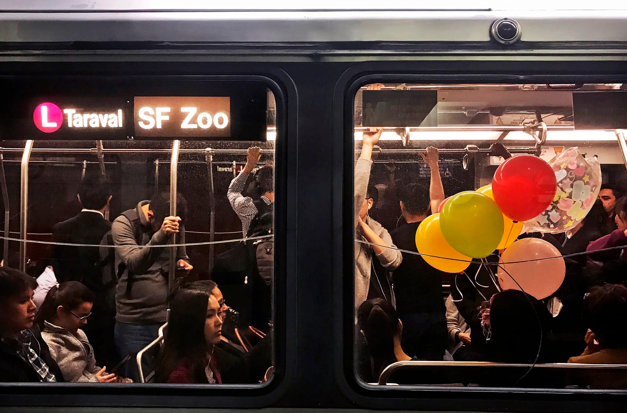 a crowded bus full of people, some holding colorful balloons, shot from the outside, with a sign that says L Taraval SF Zoo