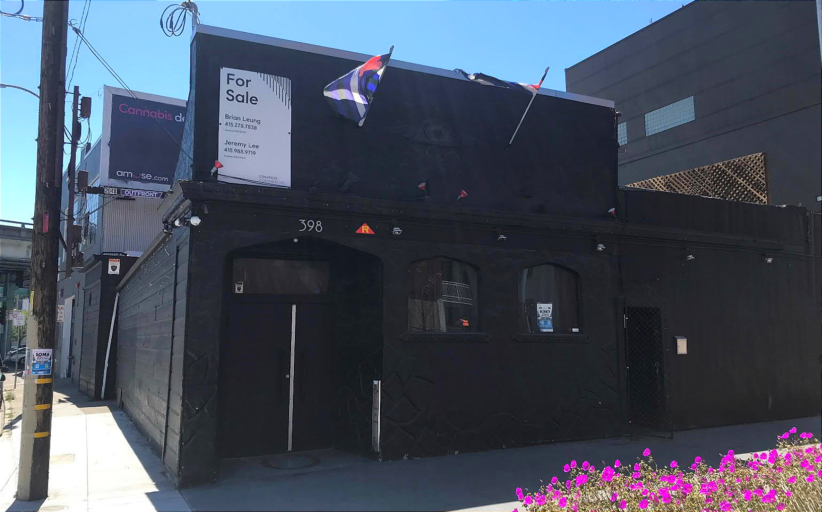 a "for sale" sign on the black exterior of a bar with purple flowers outside