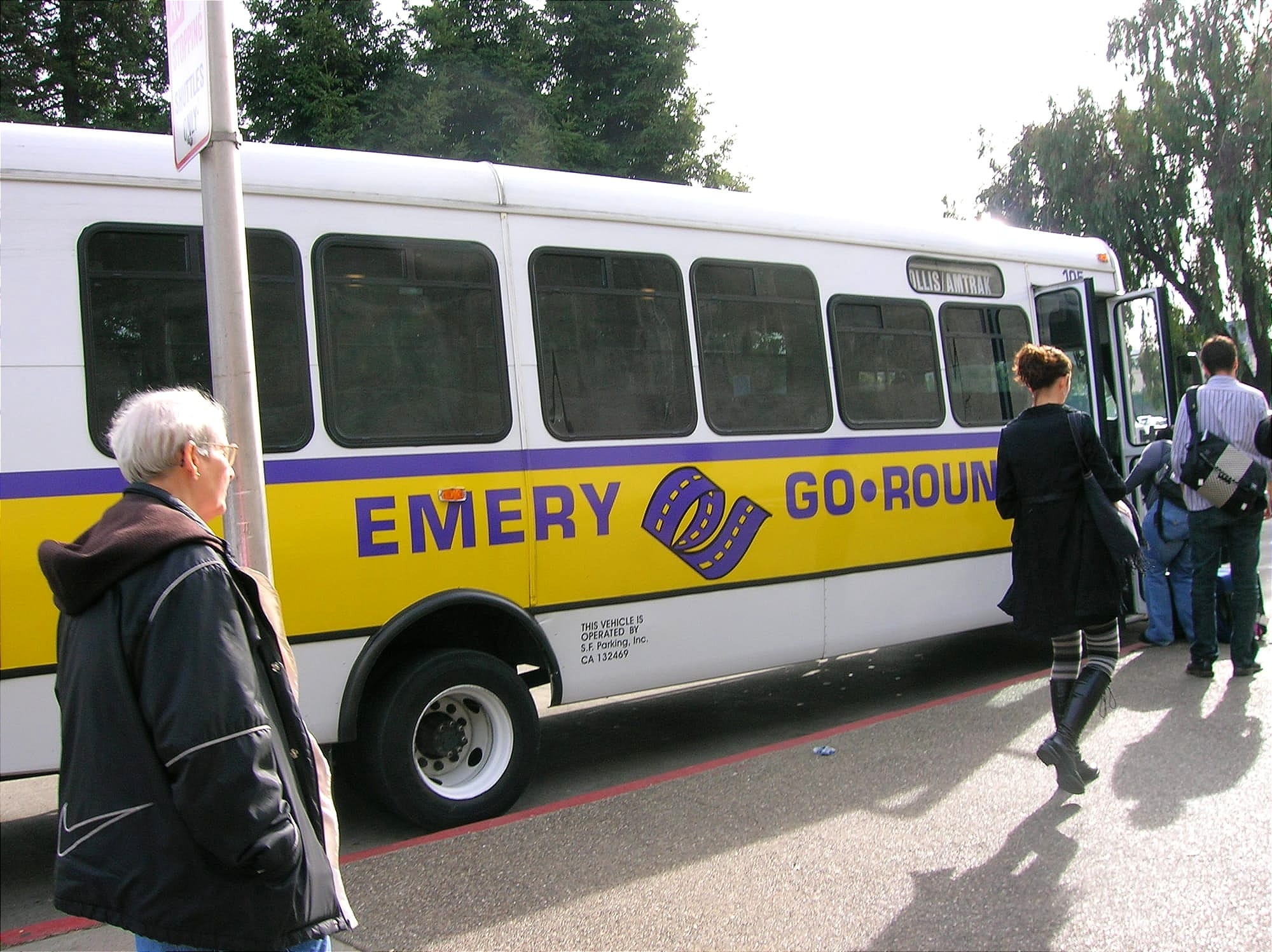 people walk to board a white and yellow shuttle bus labeled "emery go round"