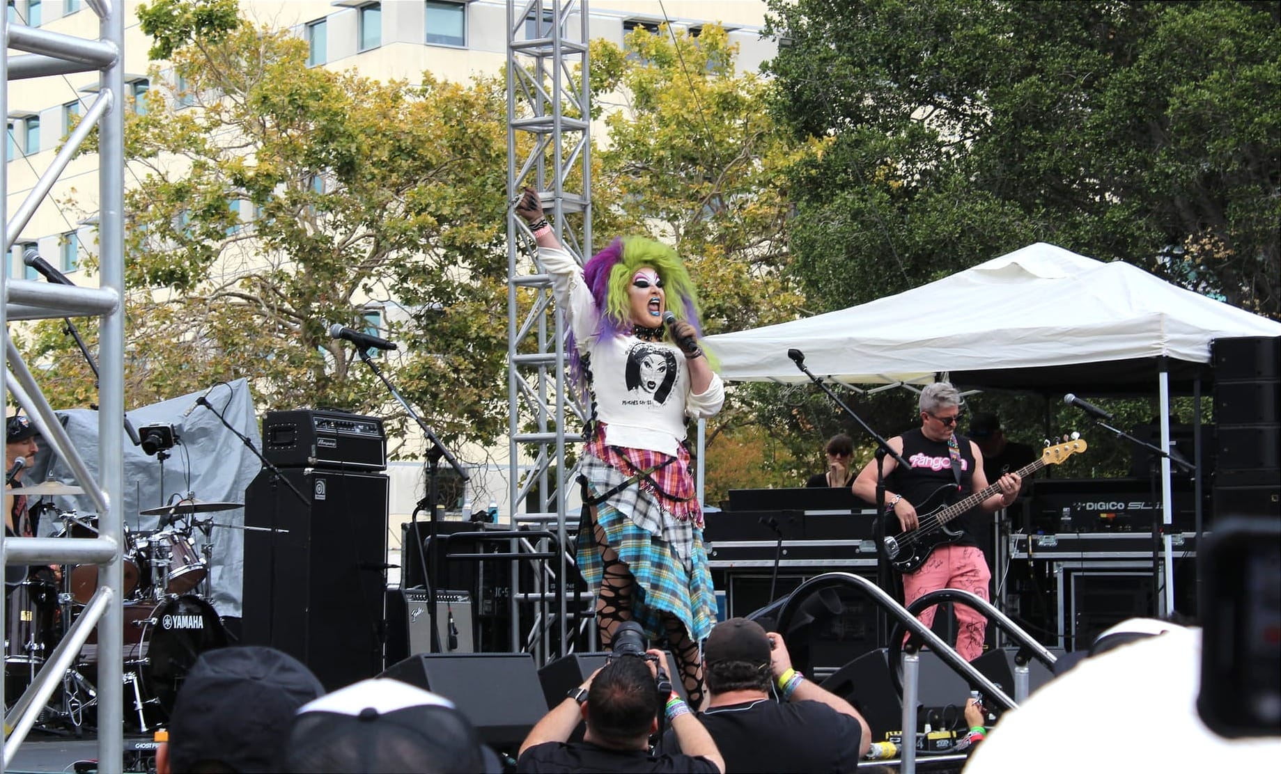 a drag performer with a green and purple wig performs on a large festival stage next to a guitarist in pink pants