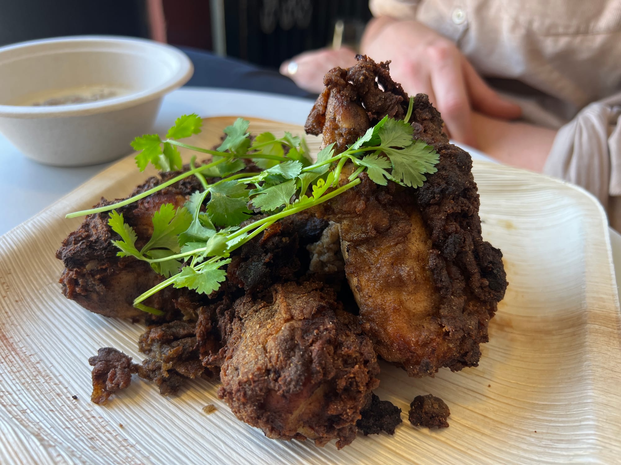 Compostable plate with four dark pieces of fried chicken topped with cilantro sprigs.