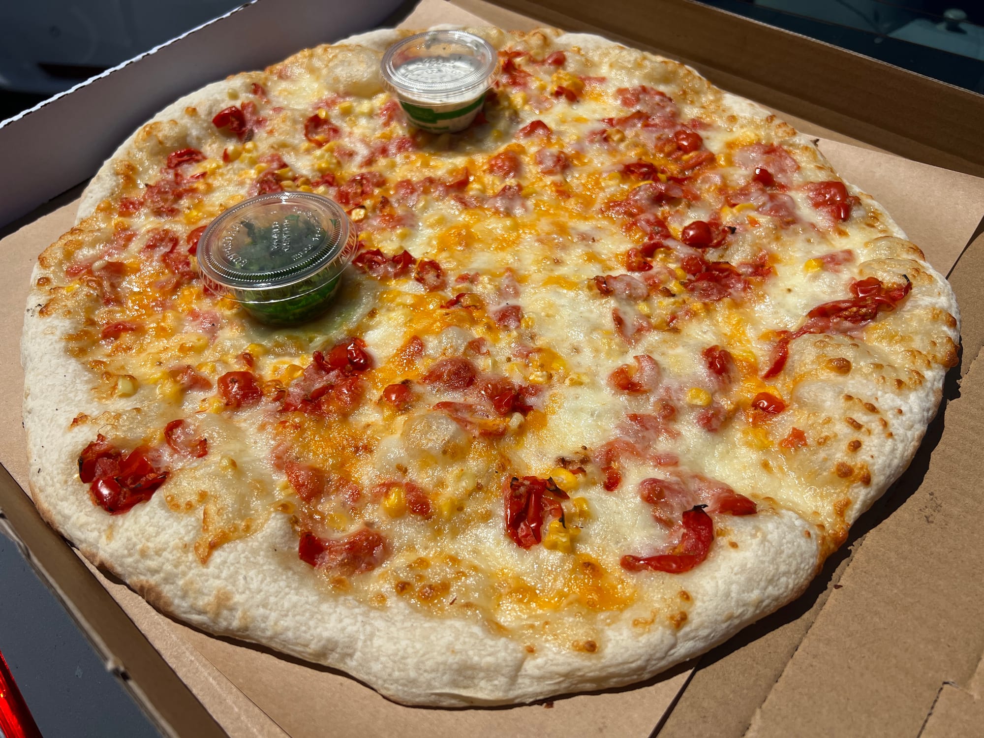 A lightly baked pizza in a box topped with corn and cherry tomatoes.