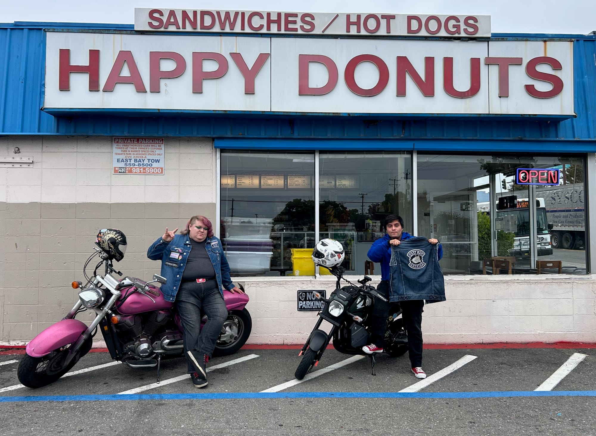 Two people stand outside a donut shop on their motorcyles. One is holding up a denim vest with a coyote patch on the back