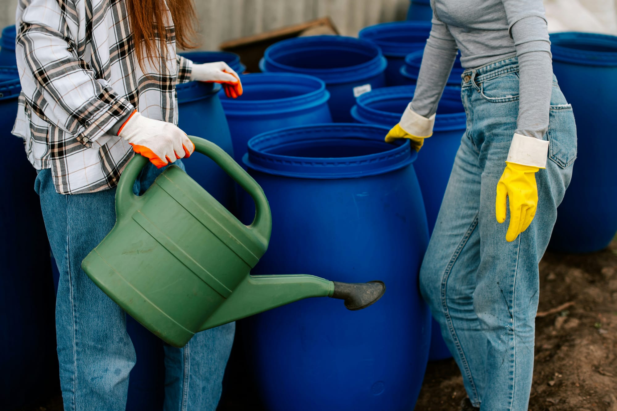 Two people stand near a bunch of blue rain barrels. One of them holds a green watering can